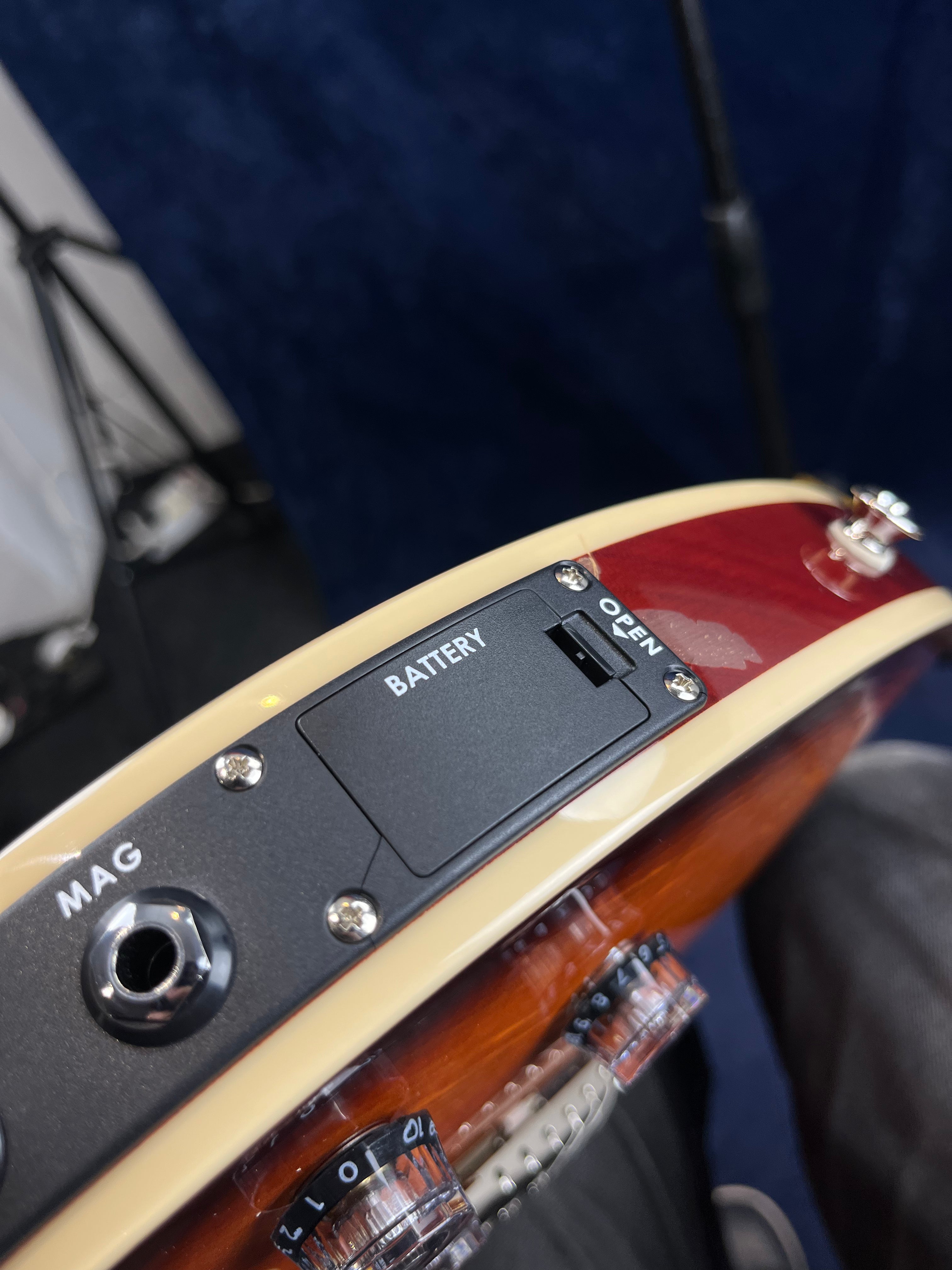 Electric guitar edge showing a MAG input jack, a battery compartment labeled "OPEN," and part of two clear control knobs.