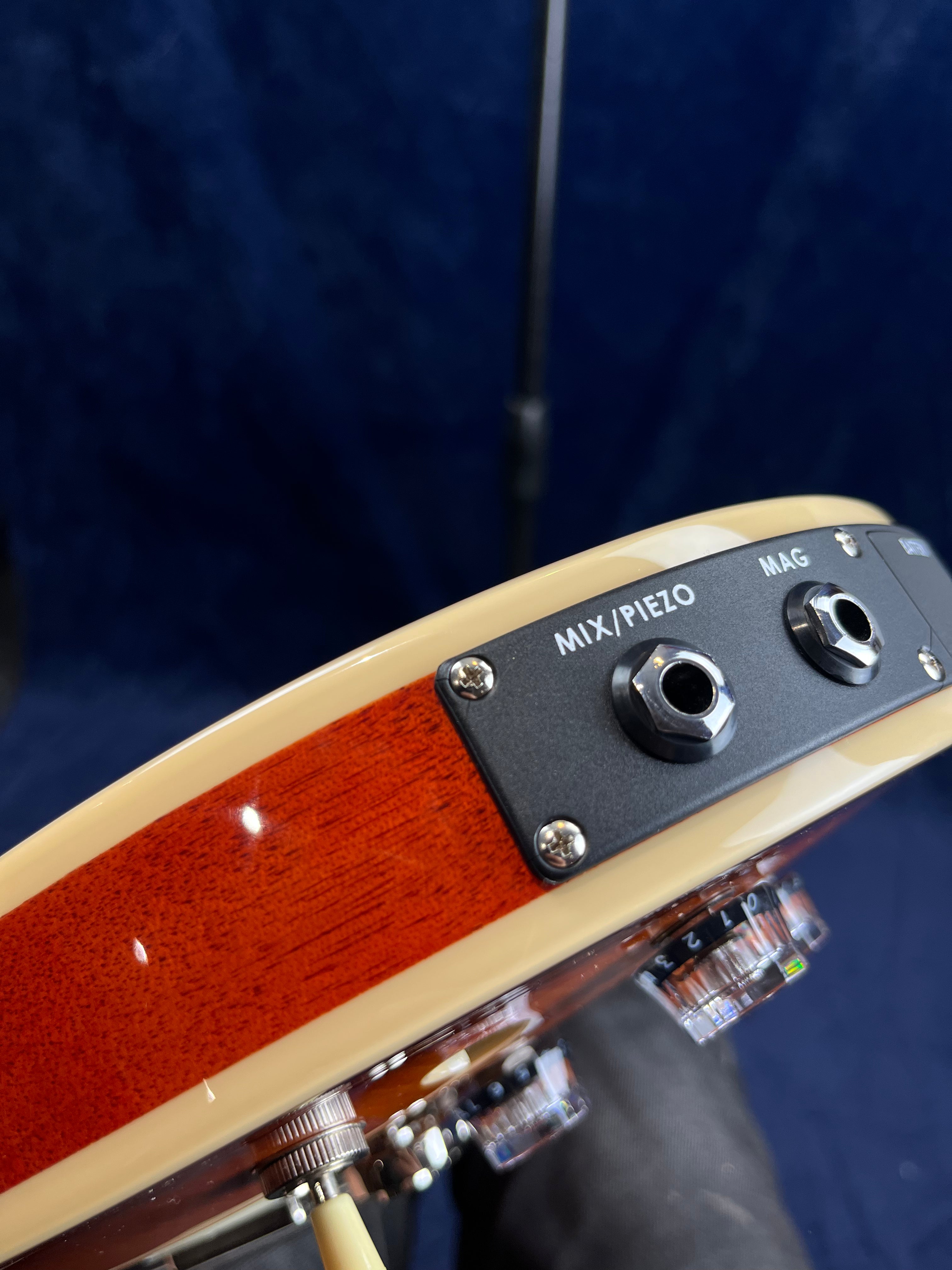 Close-up of an electric guitar’s side showing MIX/PIEZO and MAG input jacks, control knobs, cream binding, and glossy wood finish.