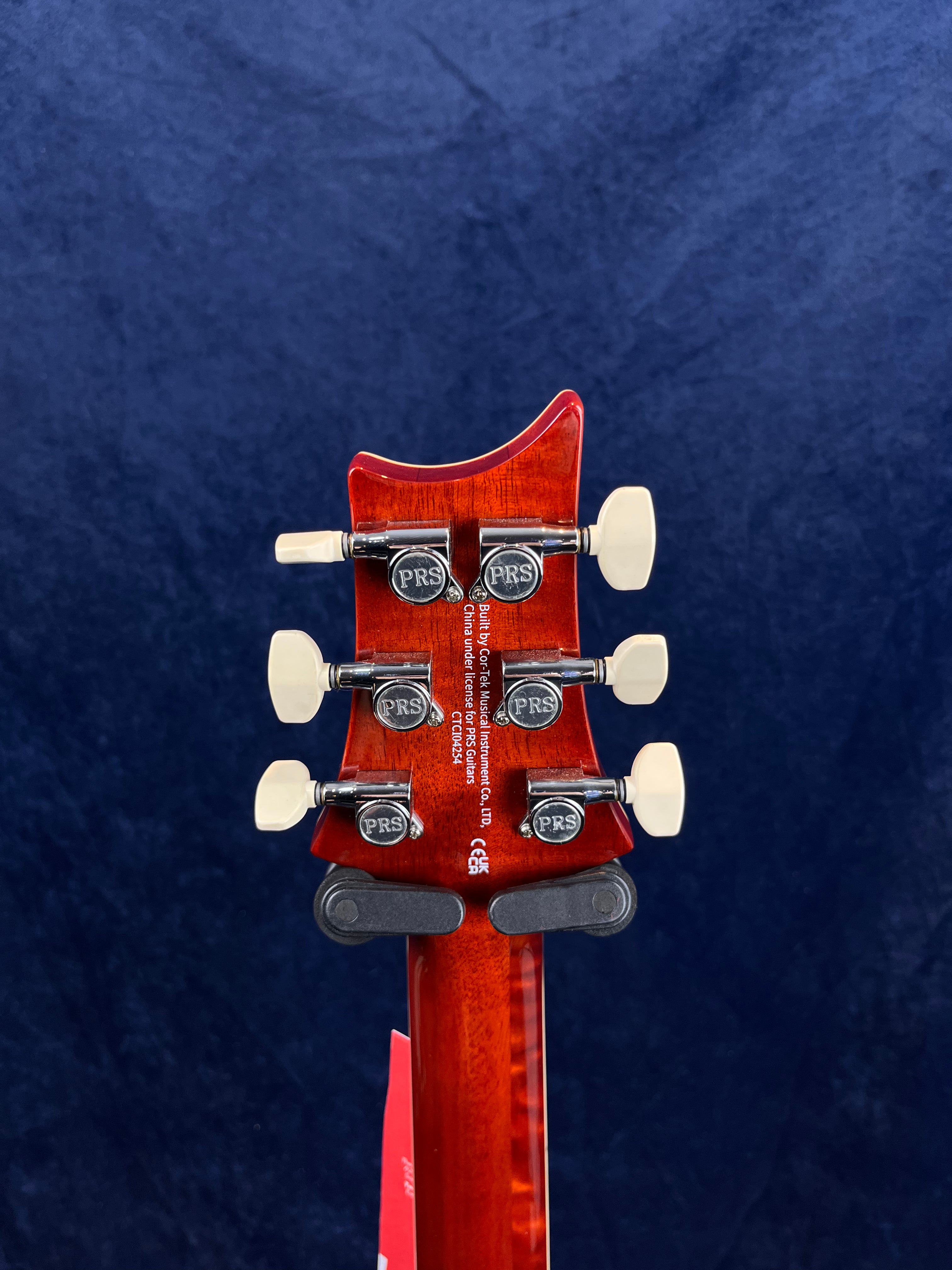 Back of a red electric guitar headstock with six PRS tuning pegs, white tuner buttons, and white manufacturer text on the wood.