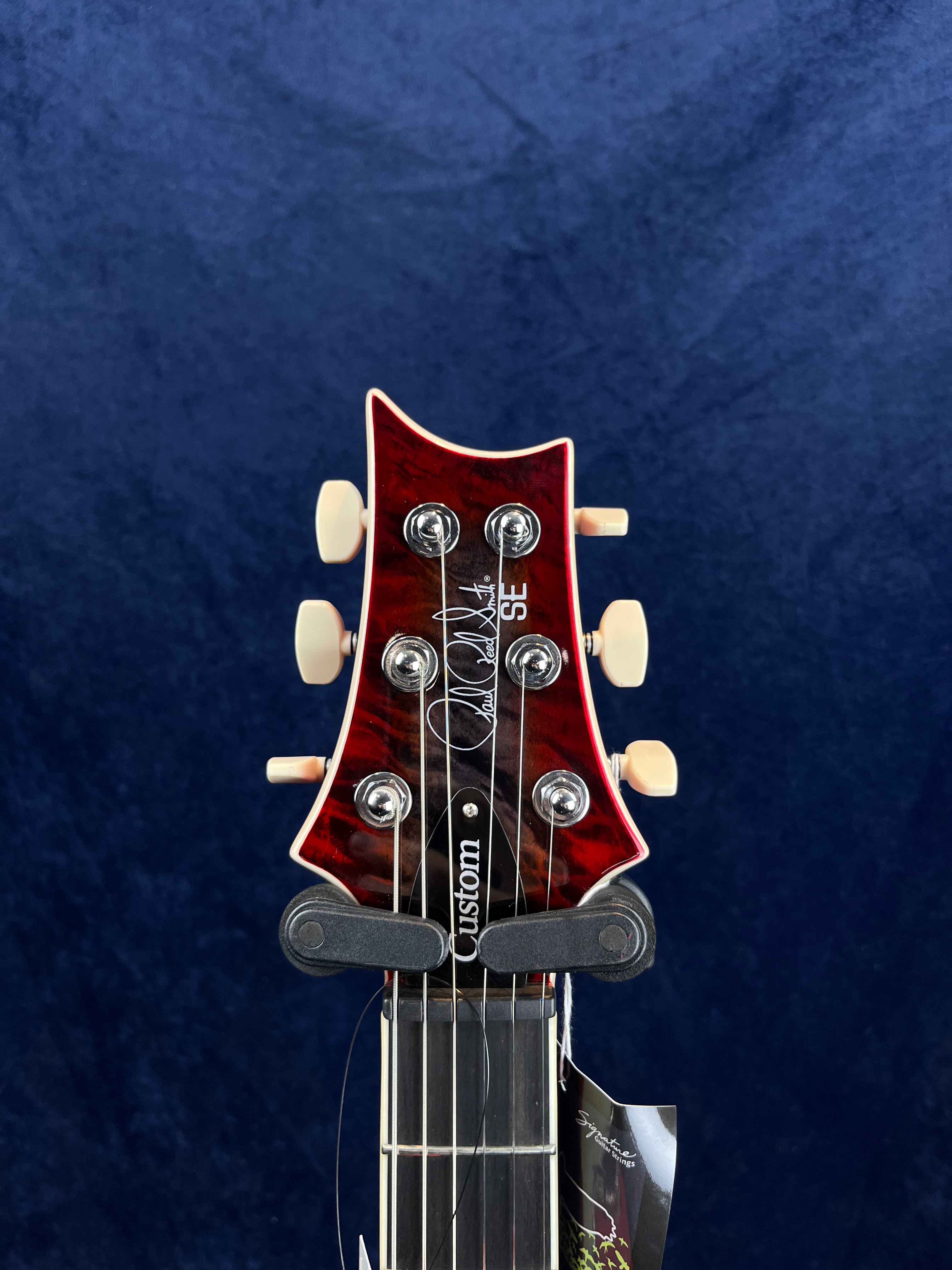 Electric guitar headstock with a red glossy finish, cream tuning pegs, and "Paul Reed Smith SE Custom" logo on dark wood, set against a dark blue background.