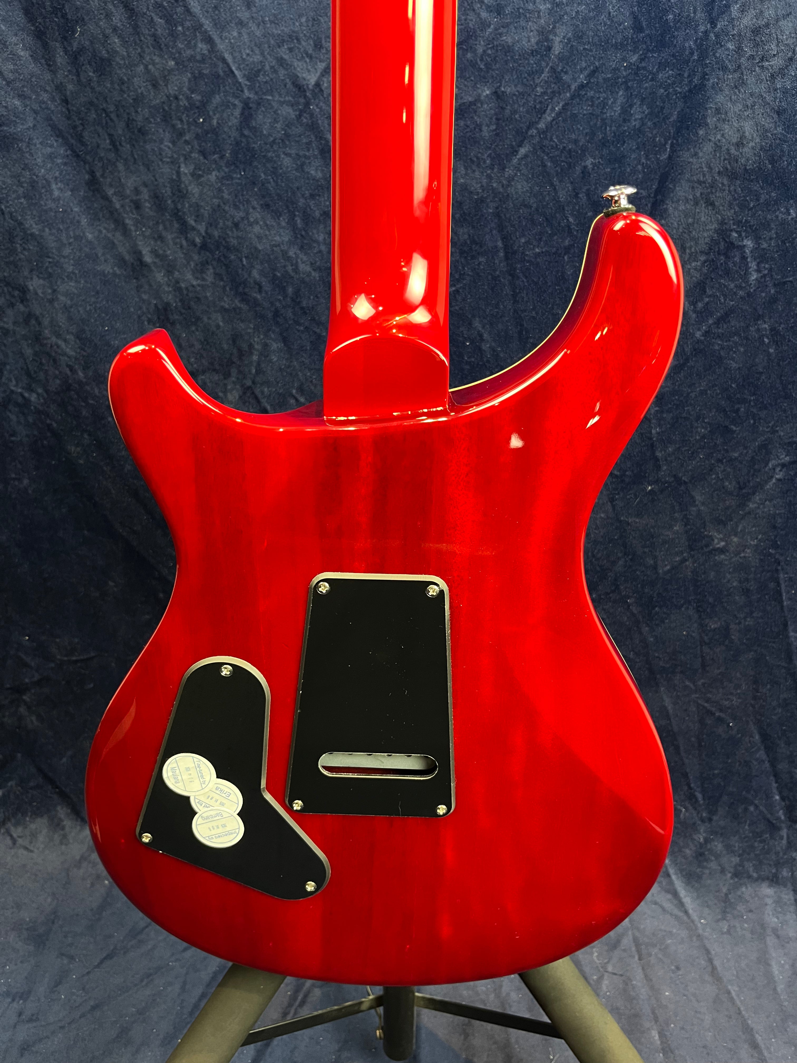 Bright red electric guitar back with black plastic cover plates, silver screw details, and a glossy finish against a dark blue background.