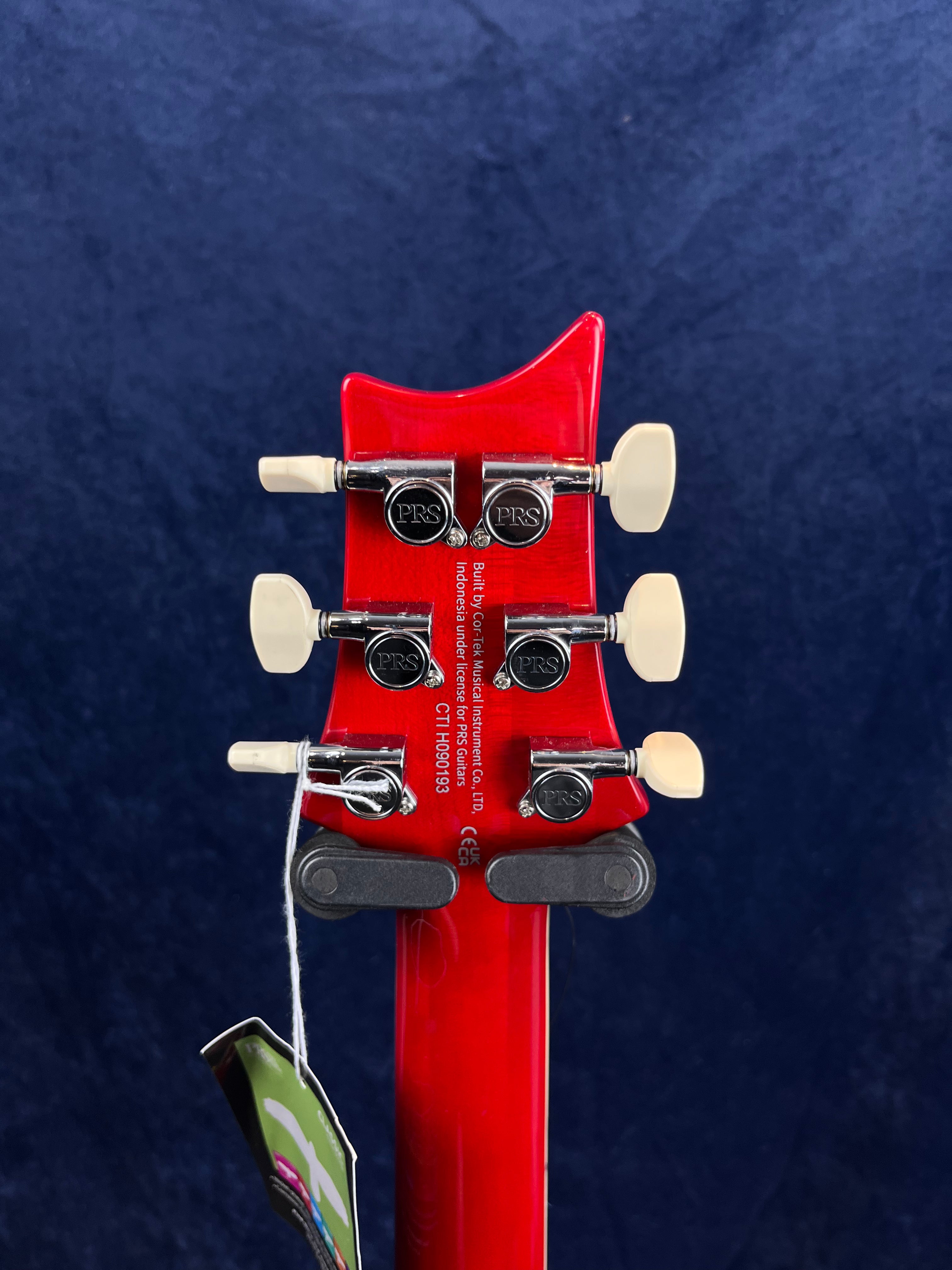 Bright red electric guitar headstock with six PRS-branded tuning pegs and white tuners, viewed from the back against a dark blue background.