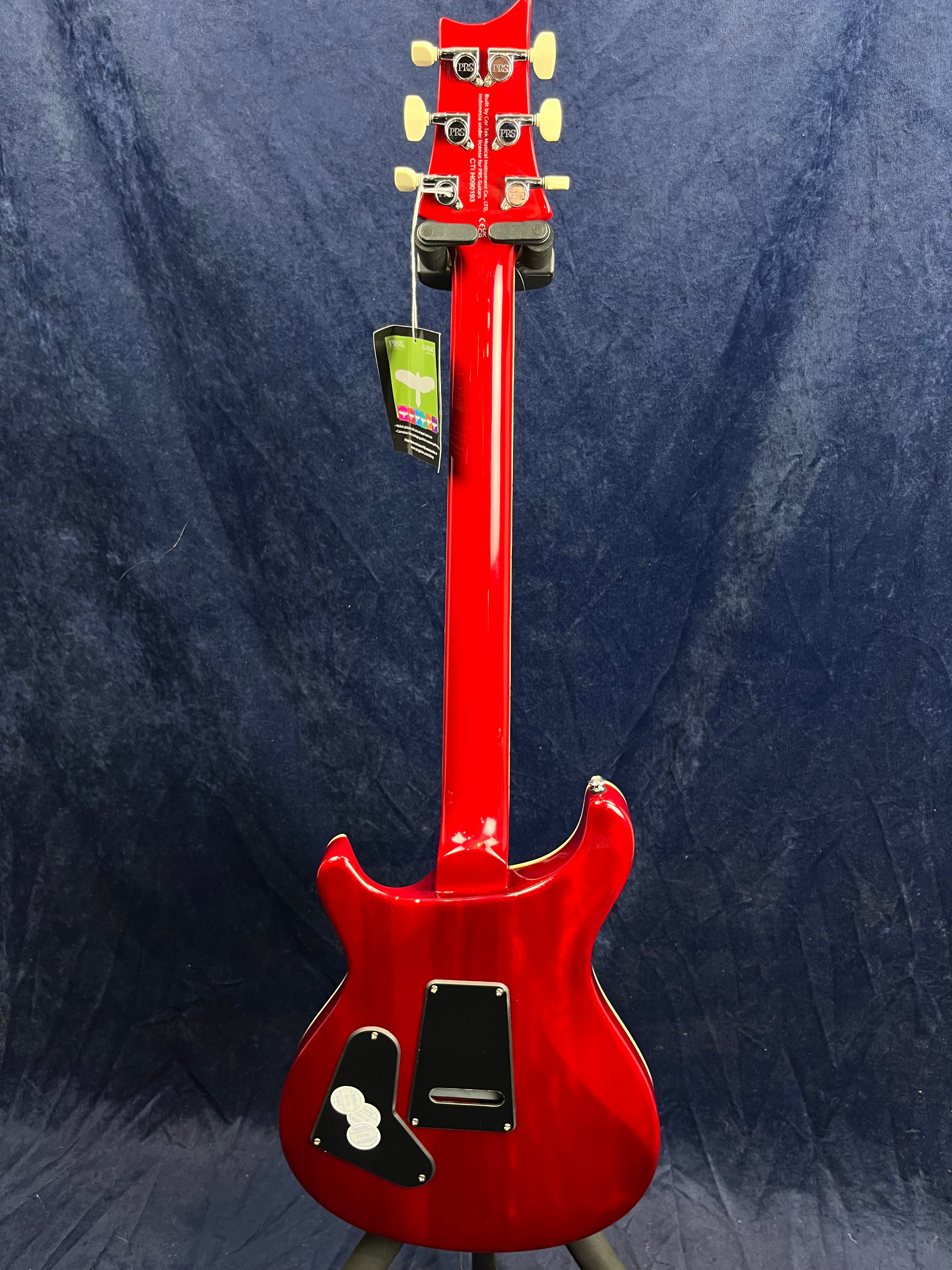 A glossy red electric guitar viewed from the back, showing cream tuners, black rear cover plates, and a tag hanging from the headstock.
