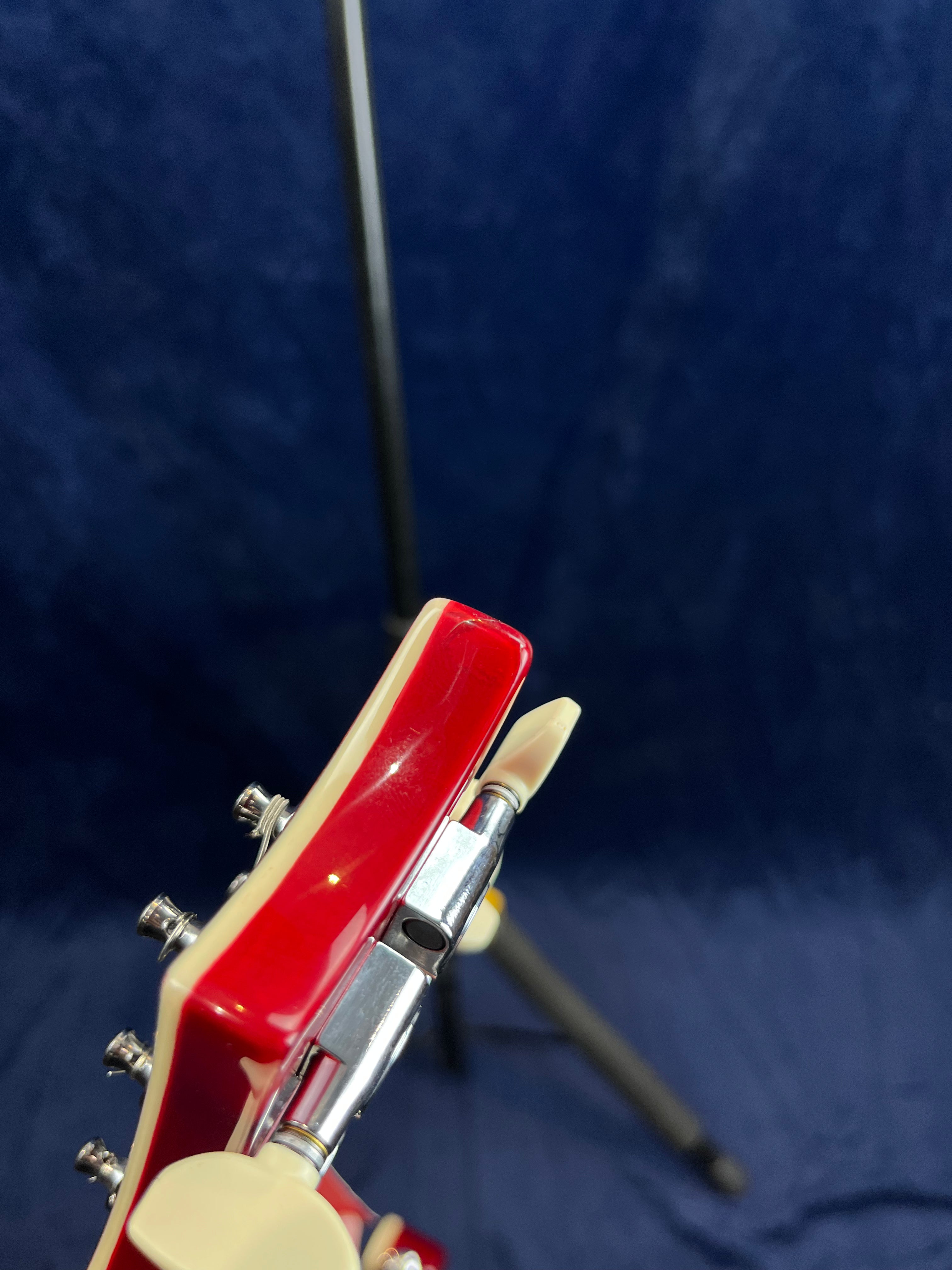 Red electric guitar headstock with cream trim, metal tuning pegs, and a silver capo clamped over the strings, blue background with tripod stand.