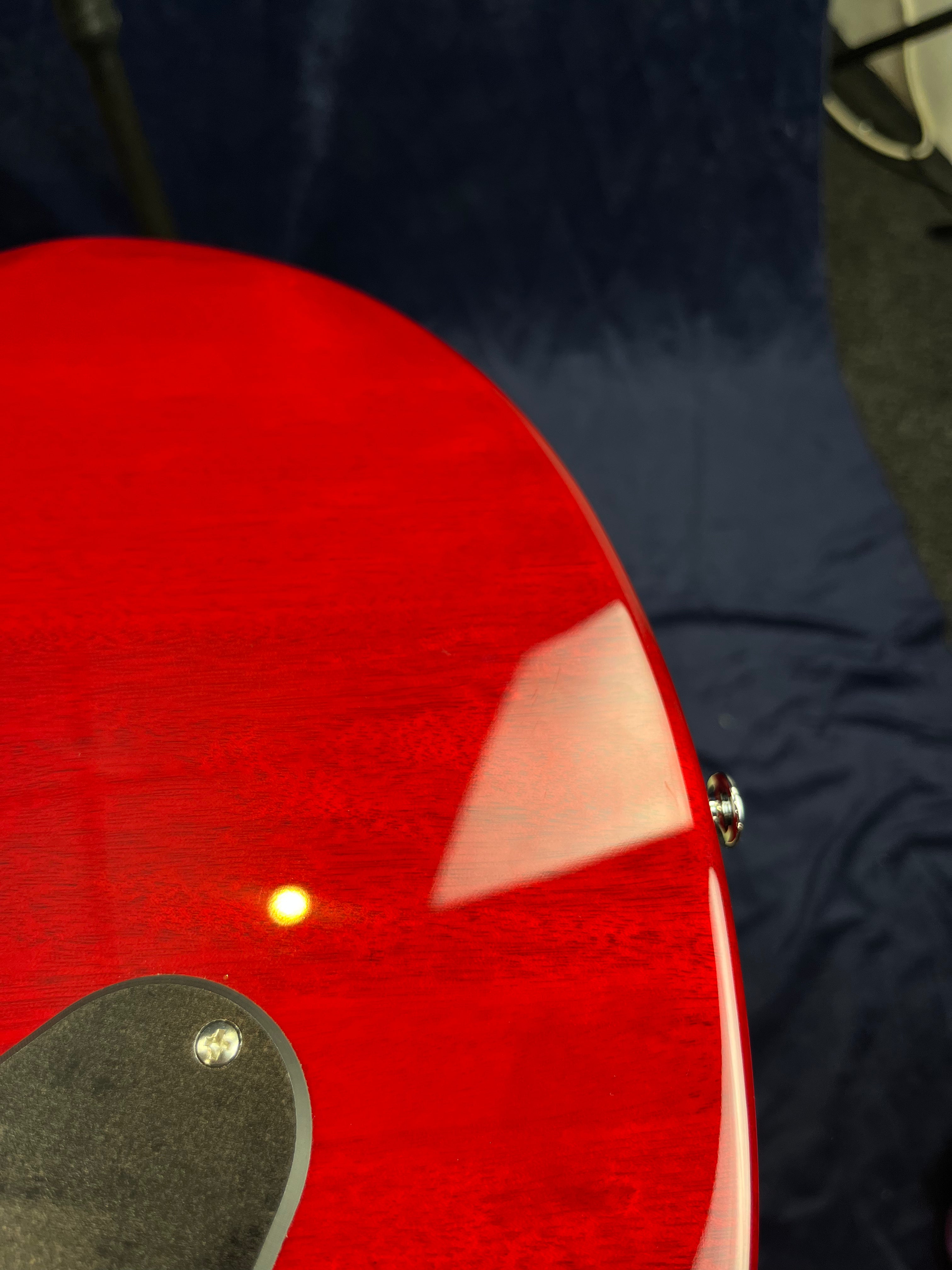 Glossy red electric guitar body with a visible control plate and screw, reflecting light and a nearby shape, set against a dark background.