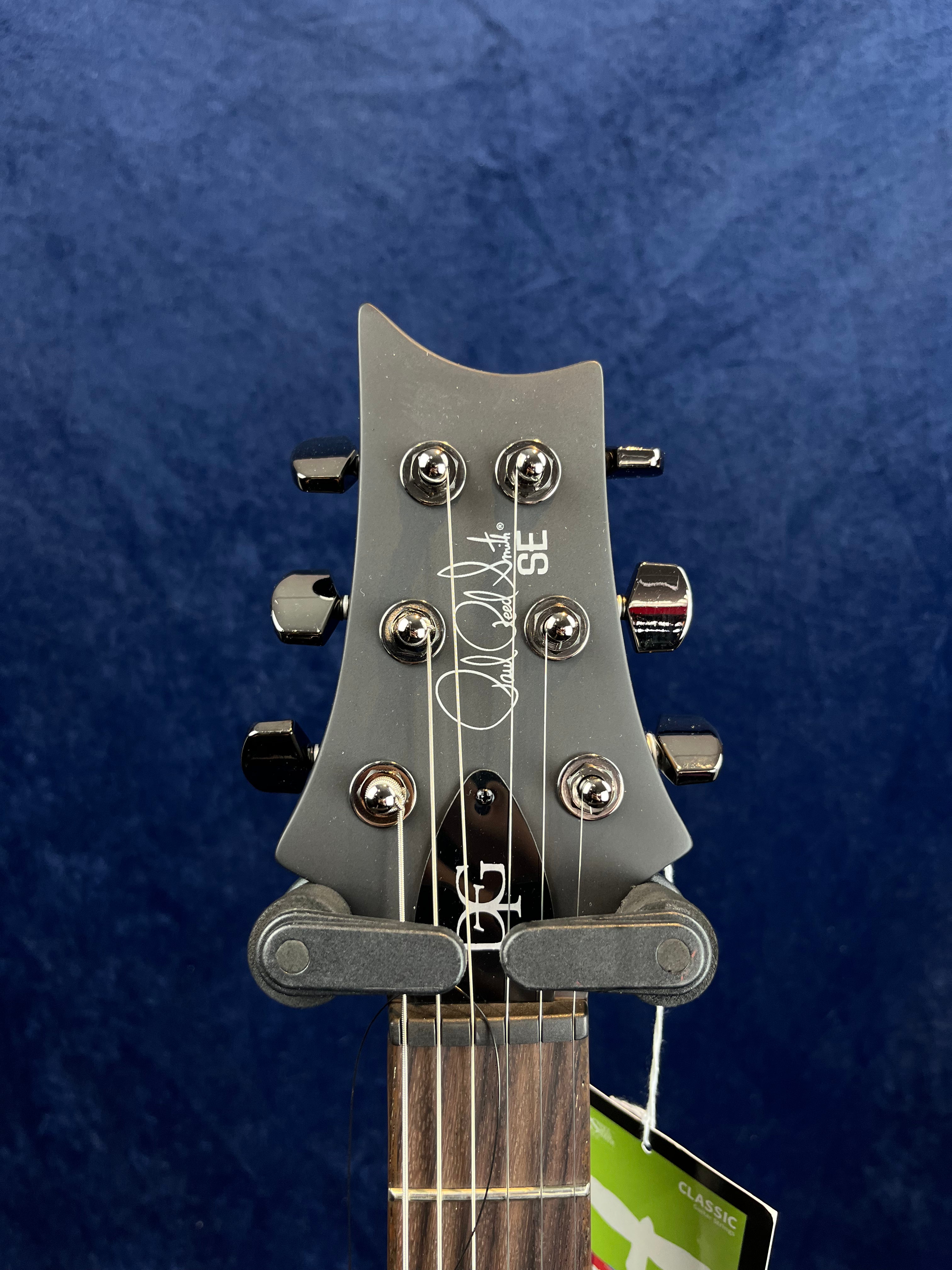 Headstock of a PRS SE electric guitar showing six tuning pegs, signature logo, and part of a guitar hanger at the nut.