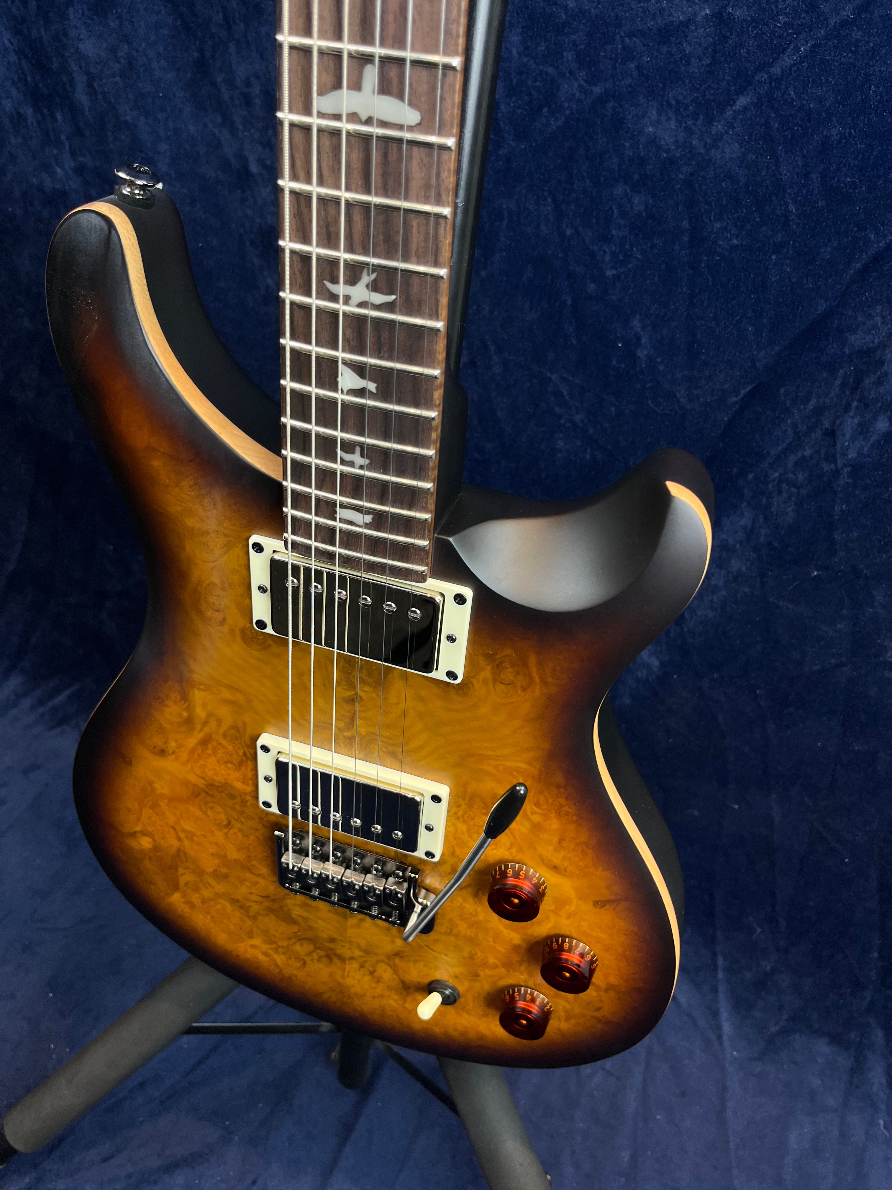 Electric guitar with a brown sunburst finish, bird inlays on the fretboard, two humbucker pickups, tremolo bar, and three amber control knobs.