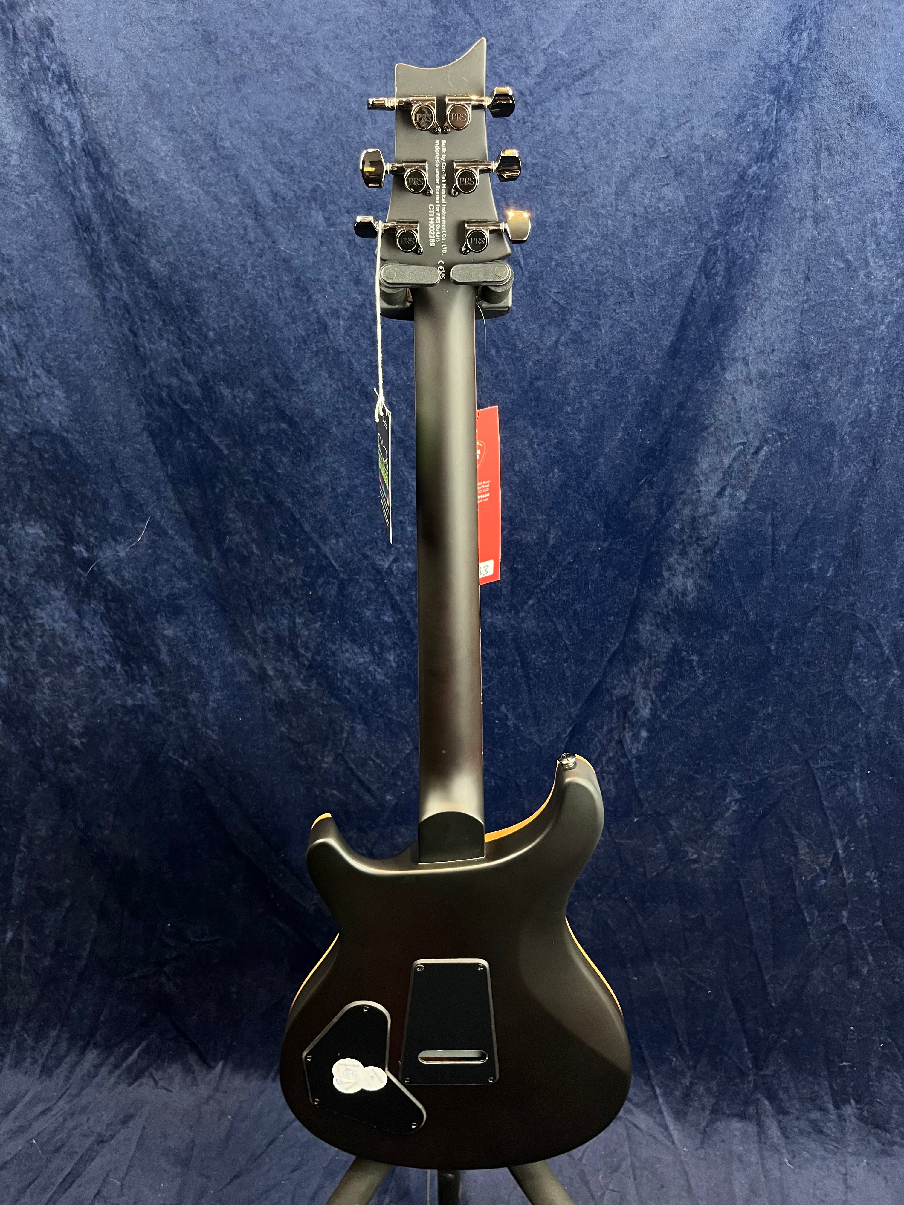 Matte black electric guitar shown from the back, flat body with two rear cavity covers, six tuning pegs, and tags hanging from the neck.