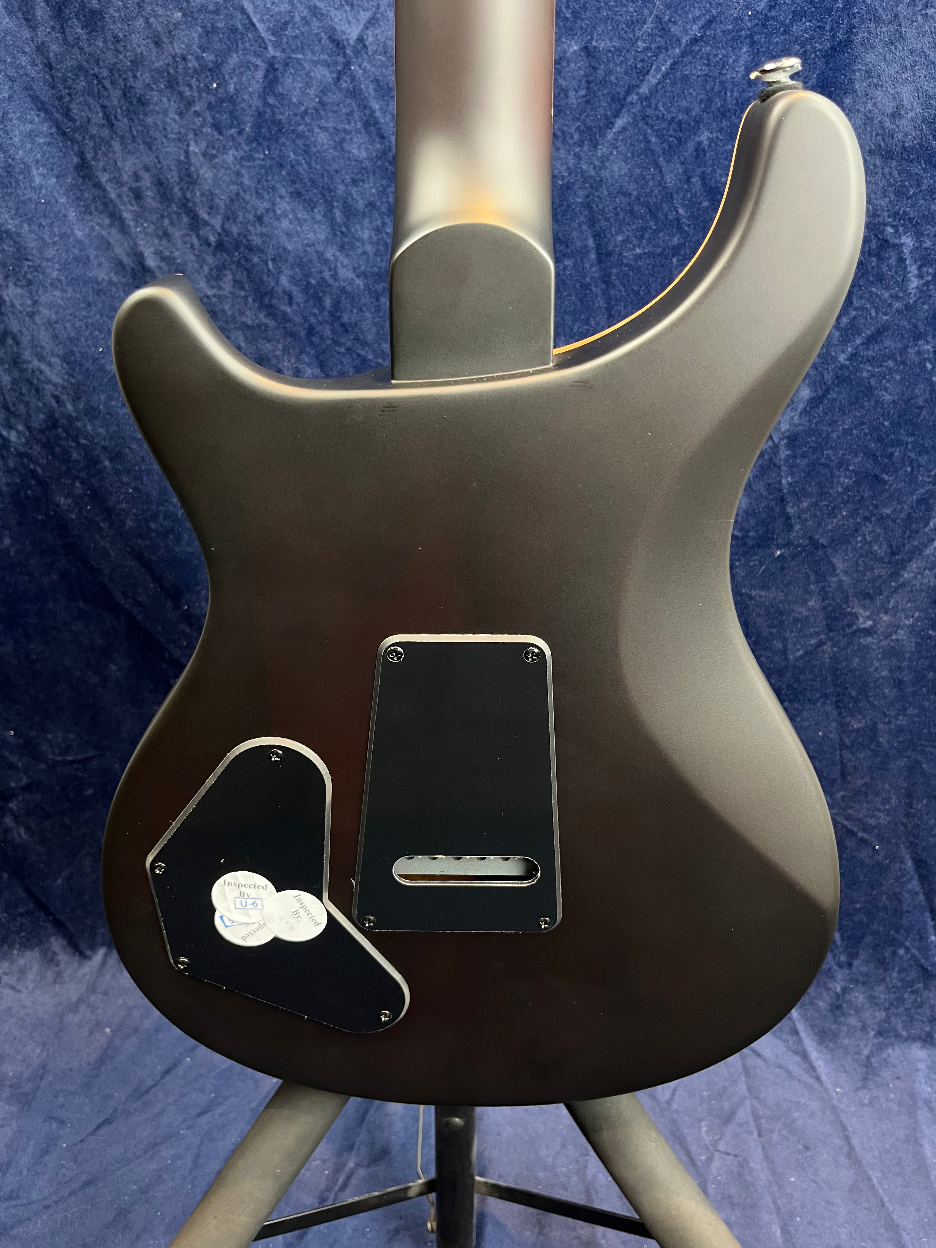 Matte black electric guitar shown from the back, with two black access panels and inspection stickers on the lower panel.