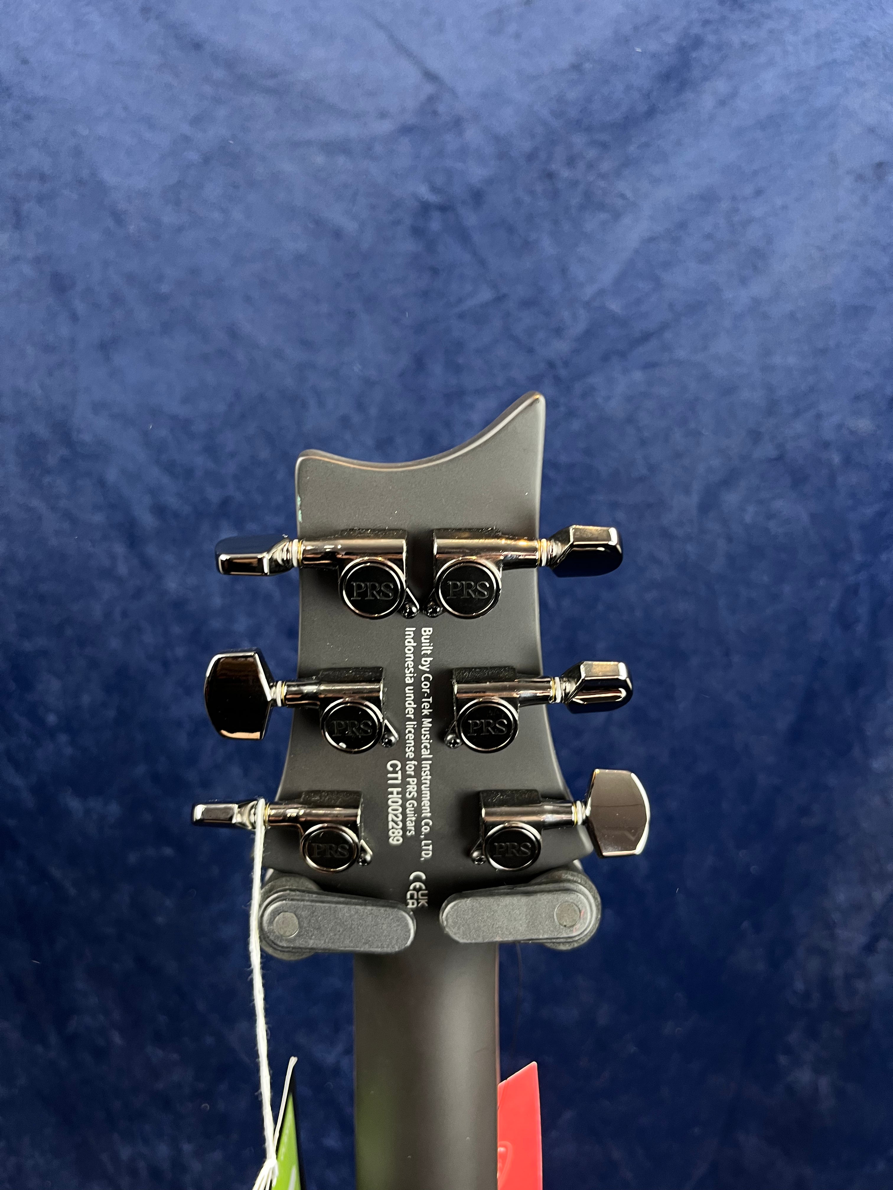 The back of a PRS electric guitar headstock shows six tuning pegs, model details, and serial number over a dark matte finish.
