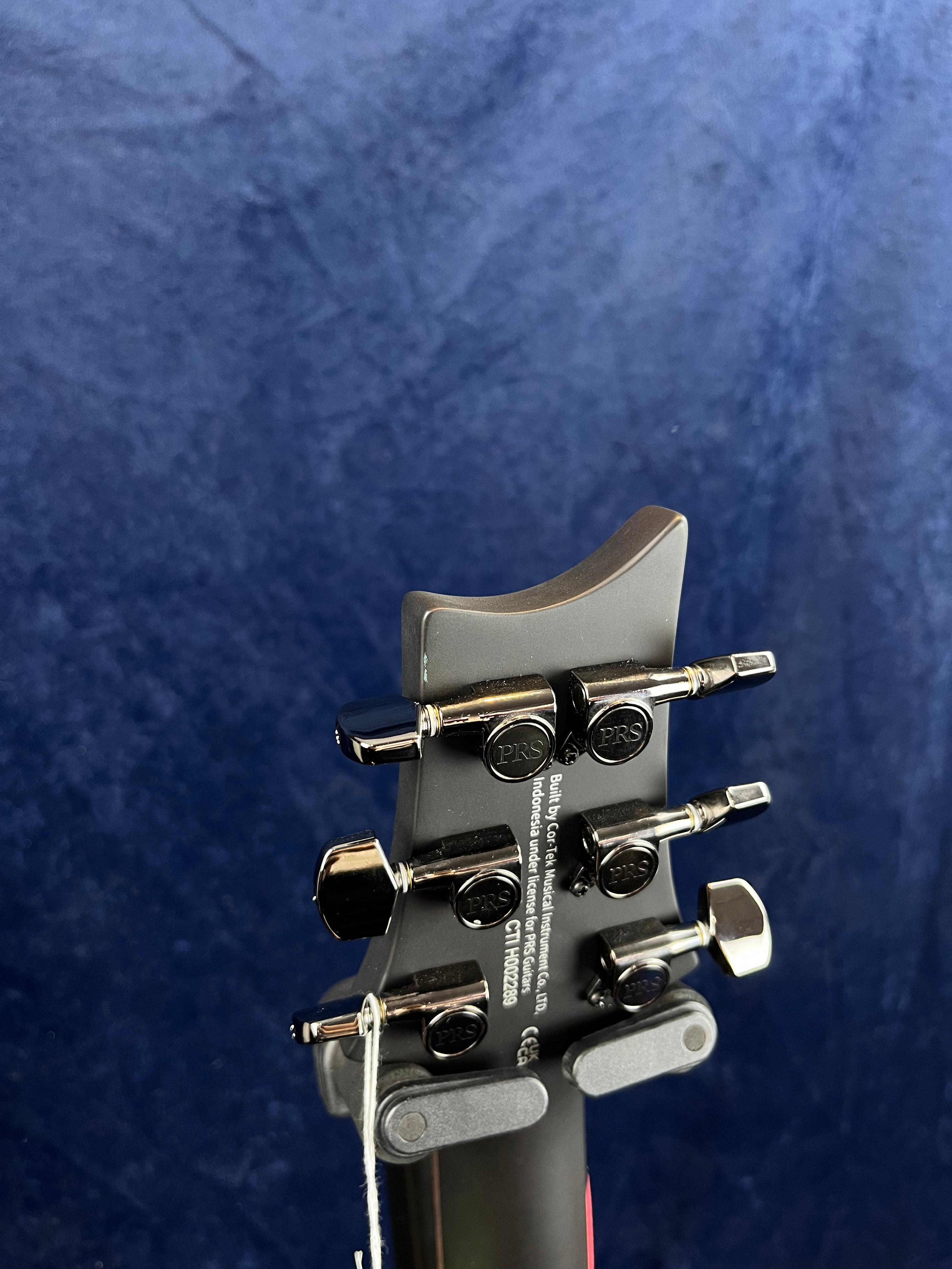 The back of an electric guitar headstock shows six PRS chrome tuning pegs, serial number, and manufacturer info against a dark blue background.