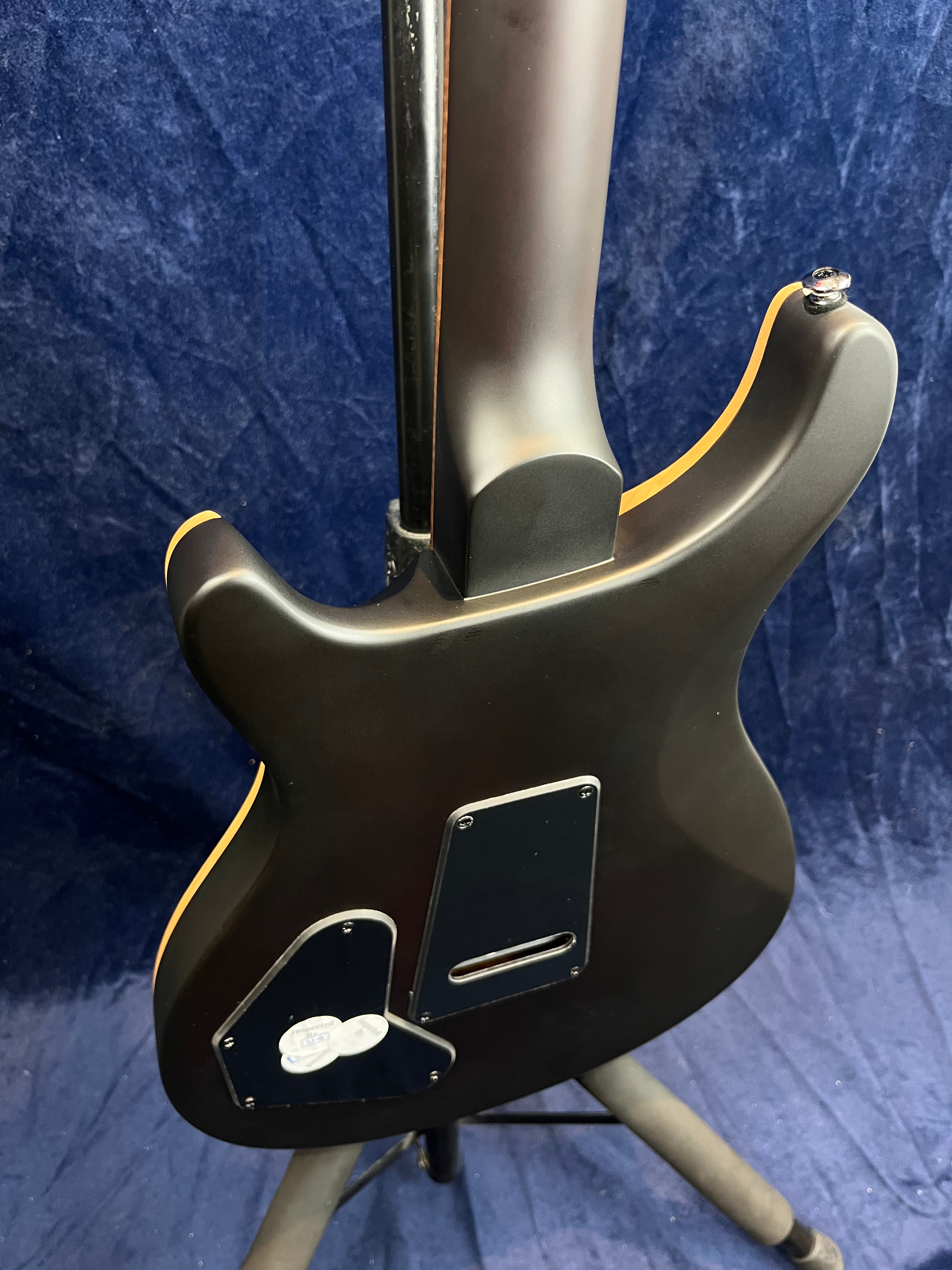 Back side of an electric guitar with a matte black finish, rear control cavity covers, and mounted on a stand against a dark blue backdrop.