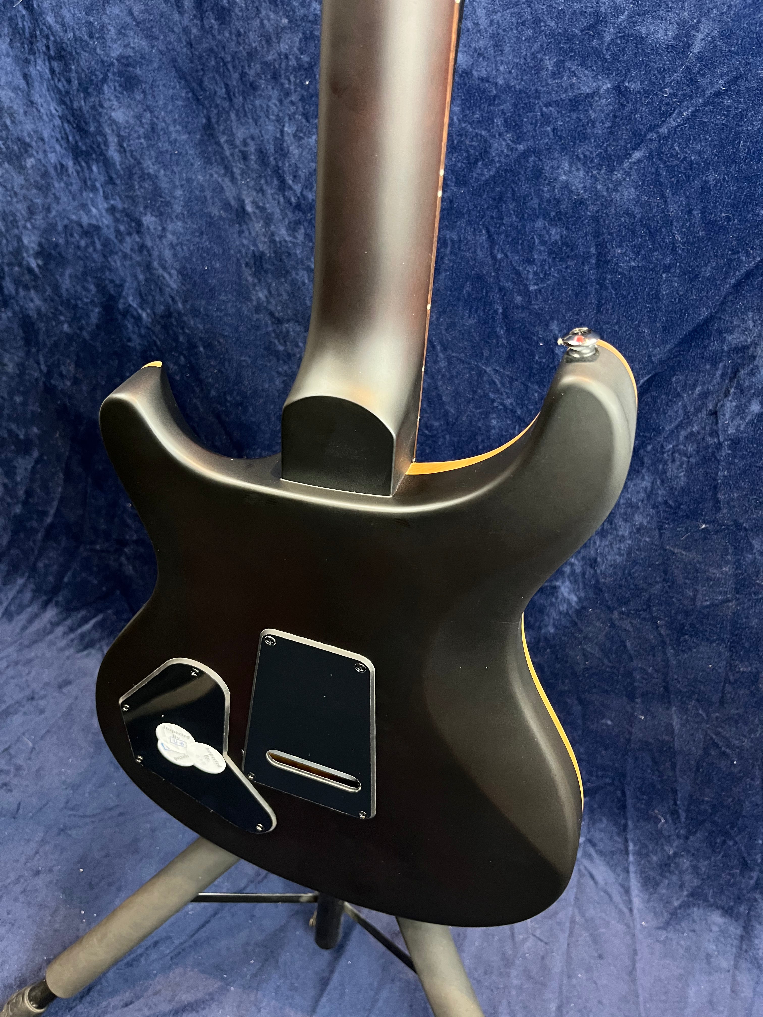 Matte black electric guitar shown from the back, highlighting the neck joint, control cavity covers, and body contours, resting on a tripod stand.