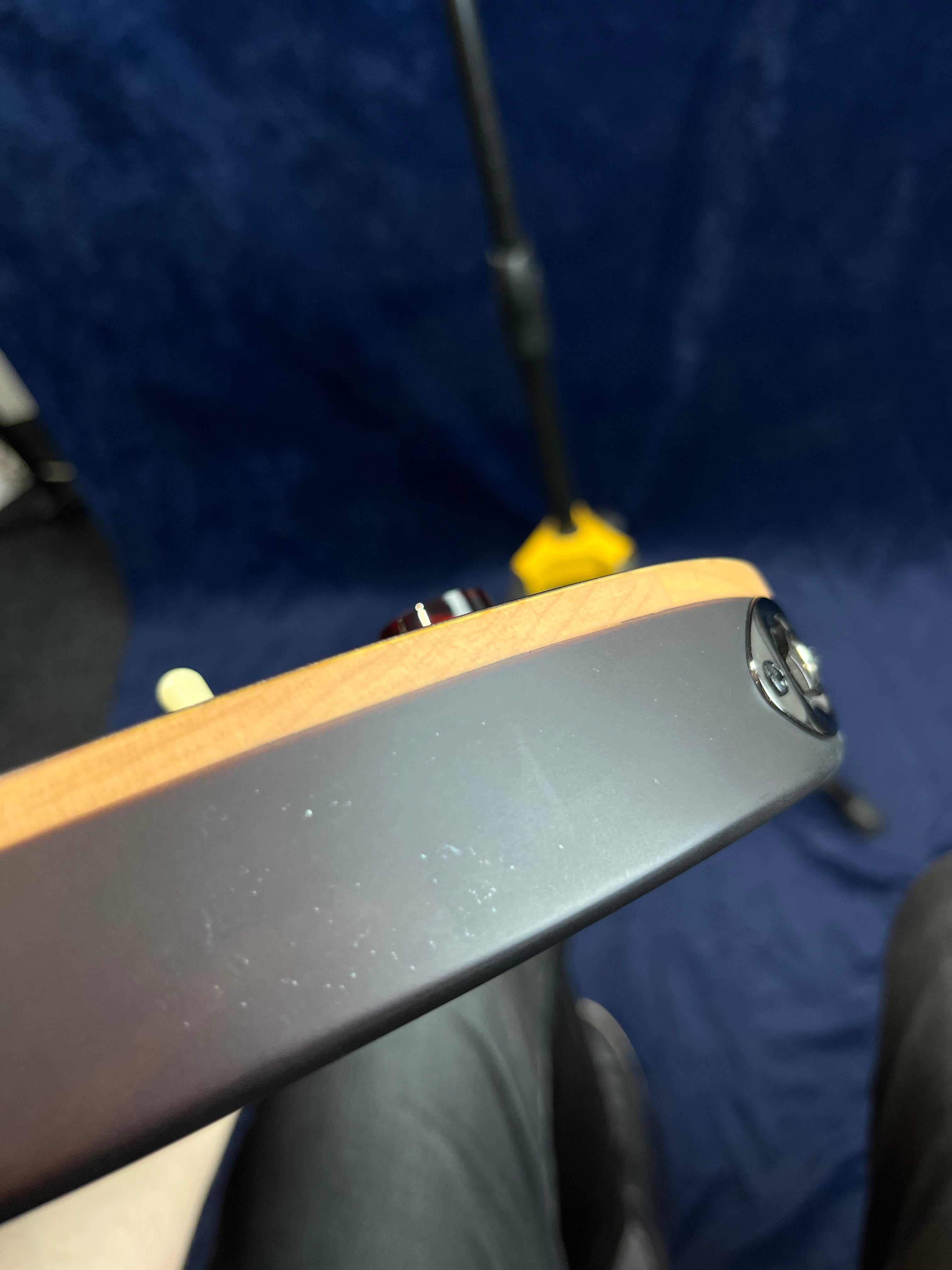 The side of an electric guitar shows minor surface scuffs on a matte black finish with a visible input jack and control knob.