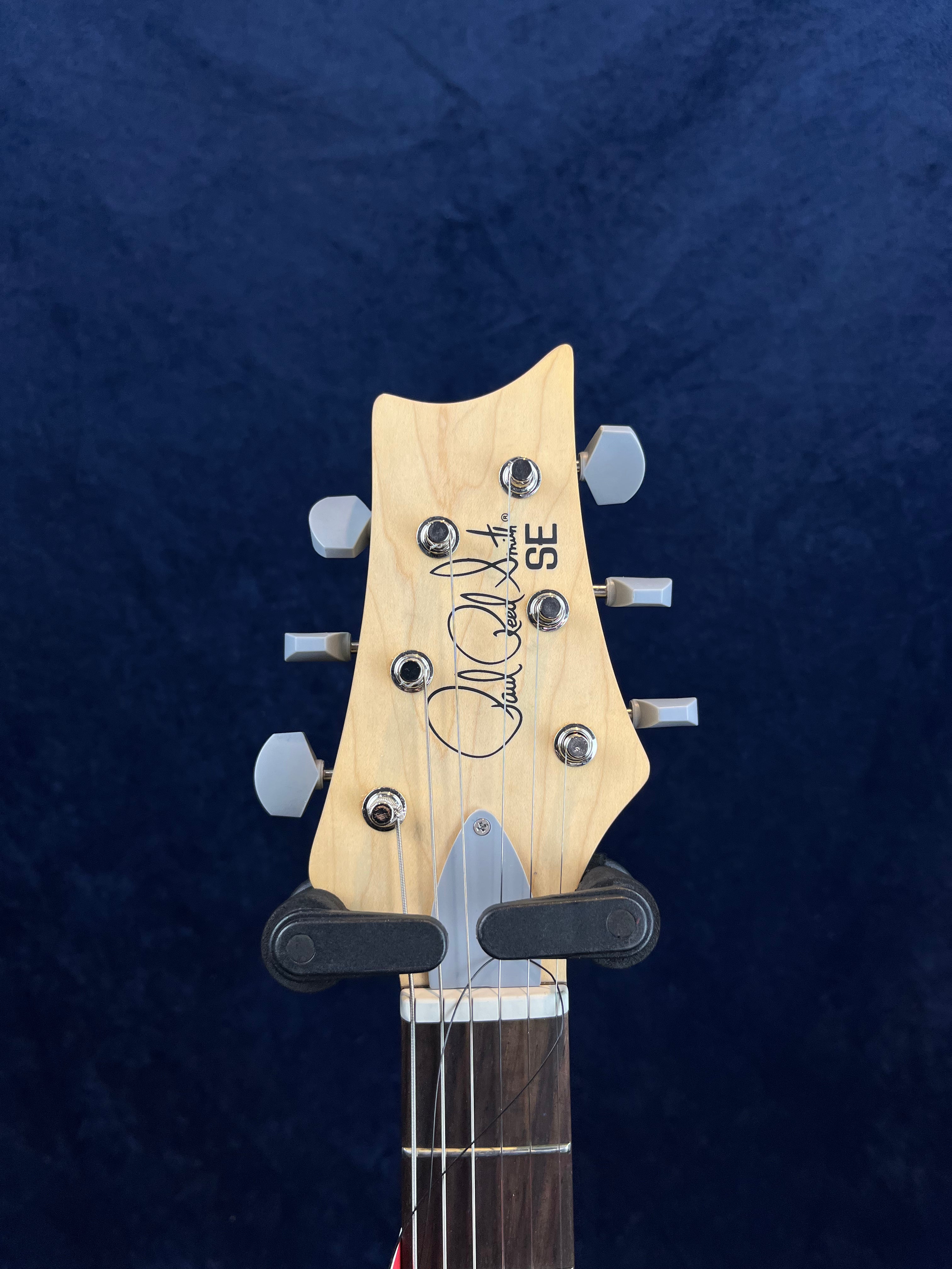Natural wood electric guitar headstock with six silver tuning pegs, black Paul Reed Smith signature, and "SE" label on a dark blue background.