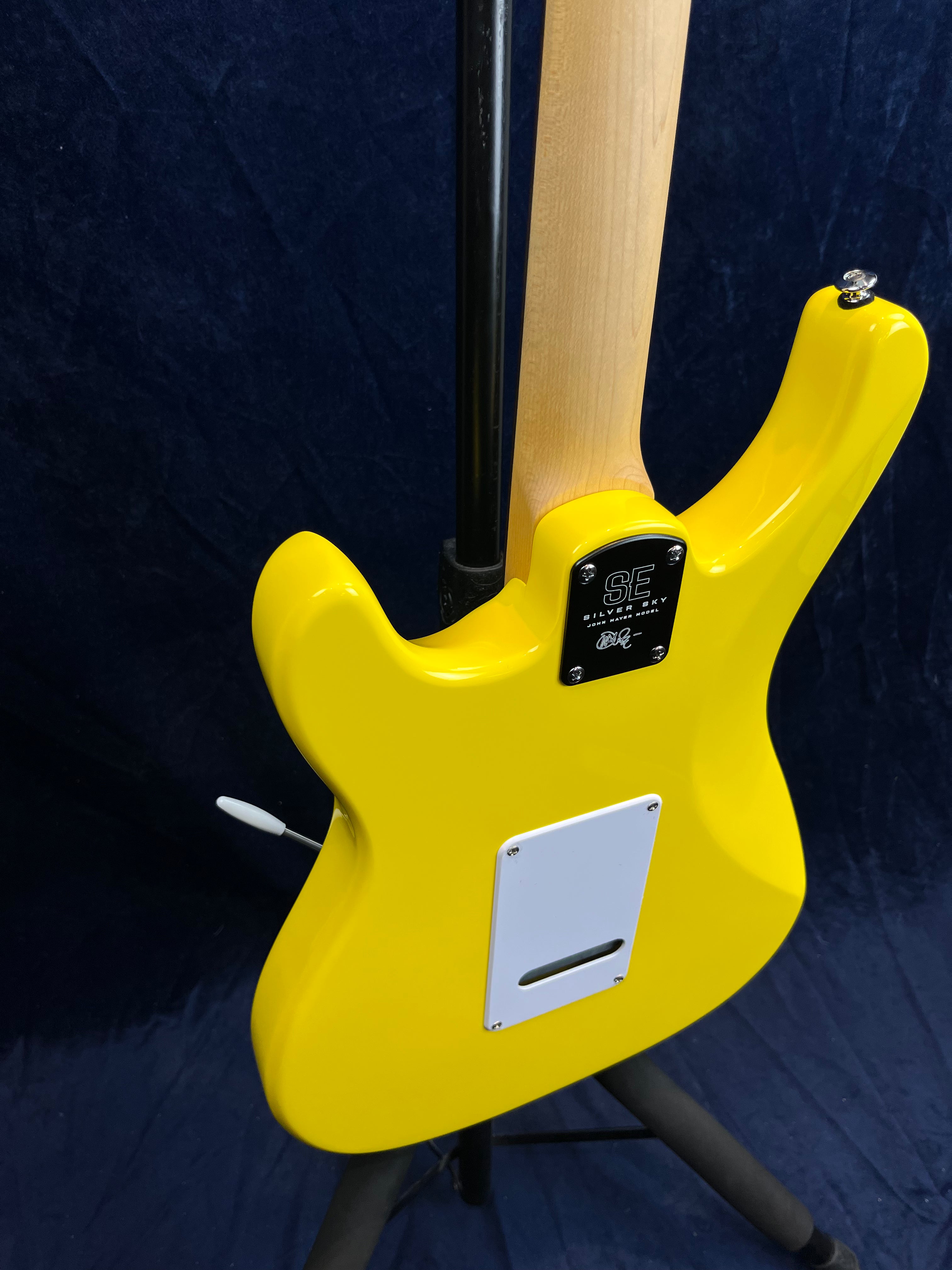 Bright yellow electric guitar seen from the back, showing the neck joint, a white backplate, and a whammy bar, standing on a tripod stand.