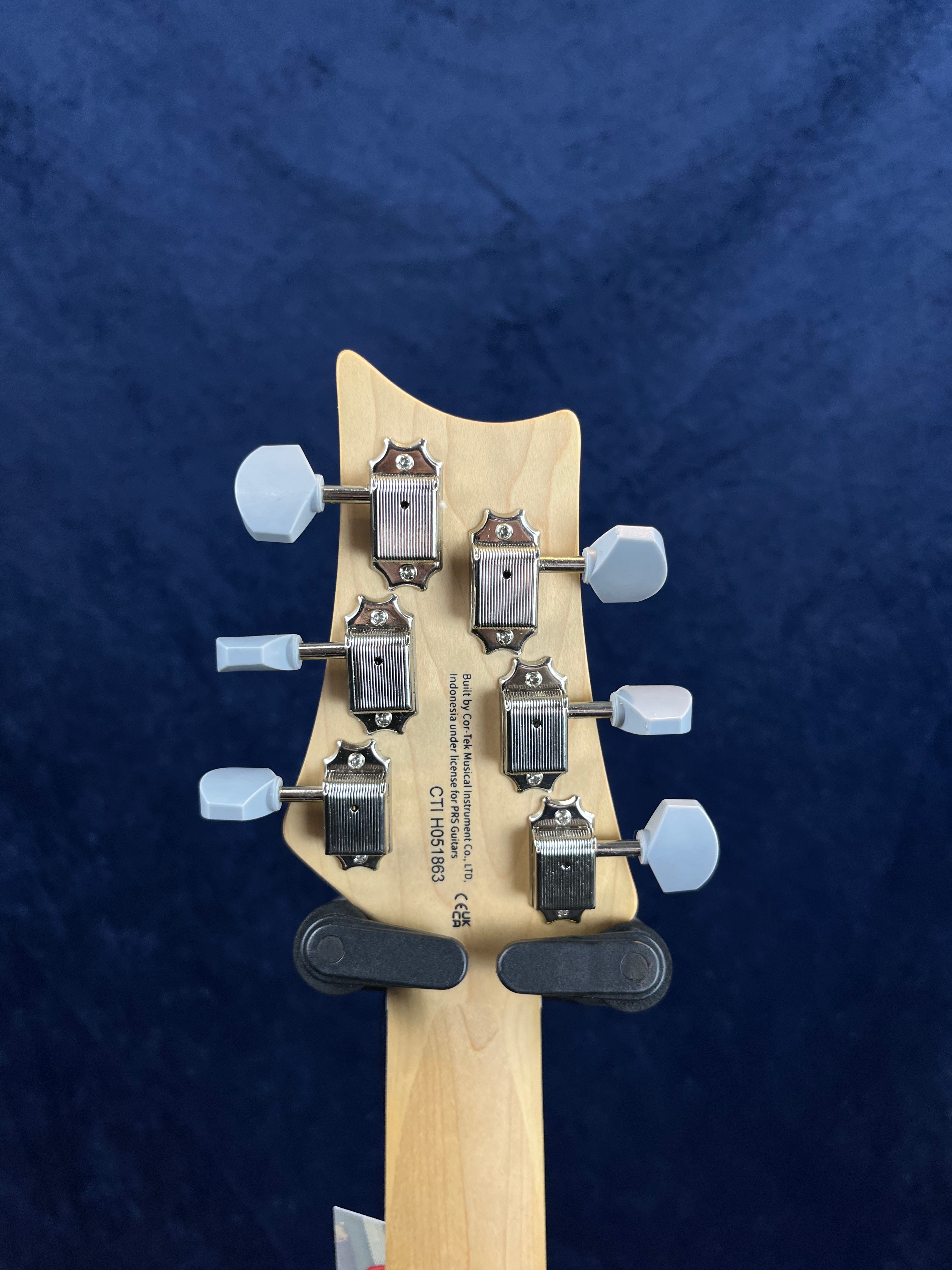 Six silver tuning pegs with white knobs on the back of a light wood electric guitar headstock, serial number and company info printed below the pegs.