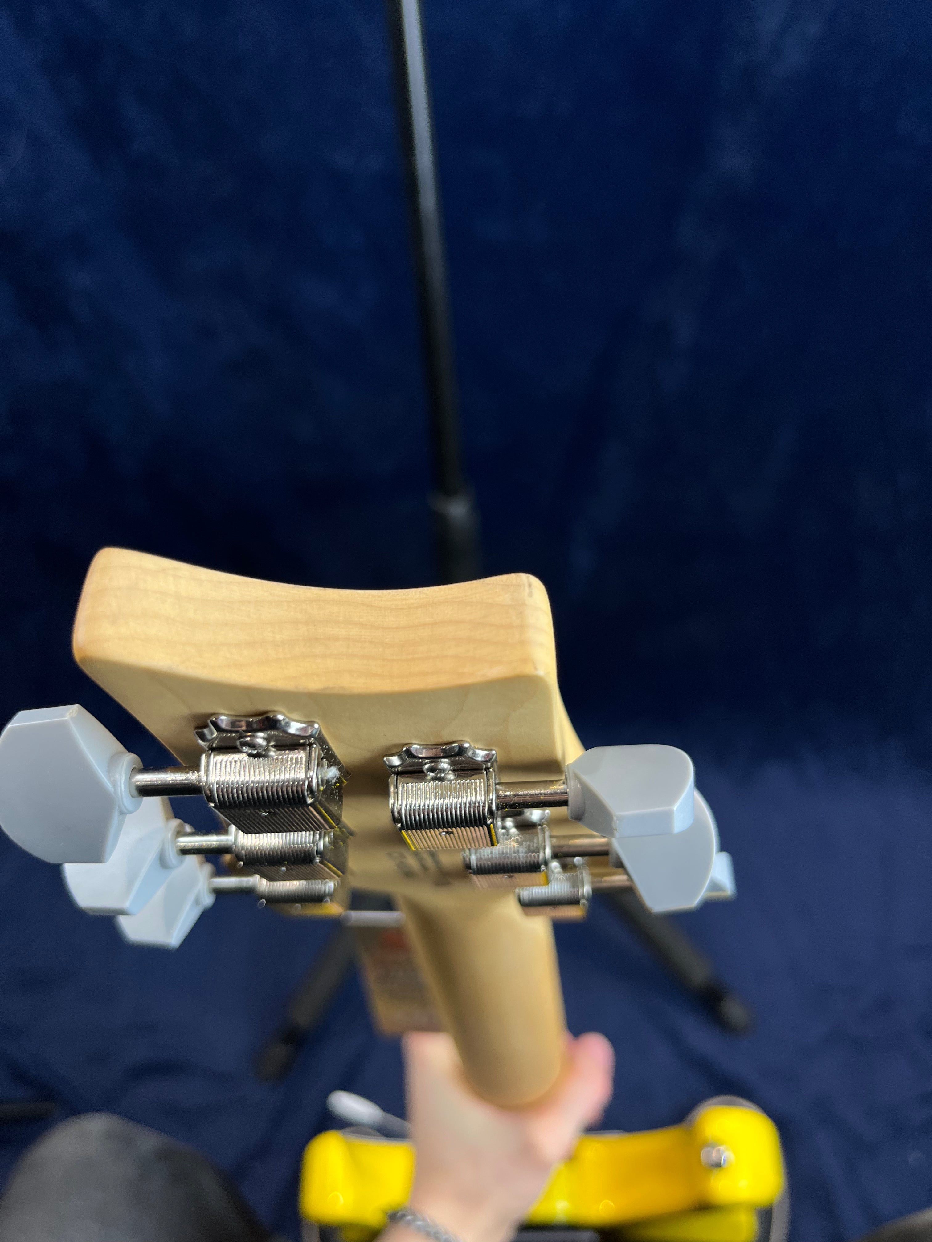 Close-up of an electric guitar’s headstock showing four tuning pegs with metal gears, held by a hand above a yellow guitar stand against a dark background.