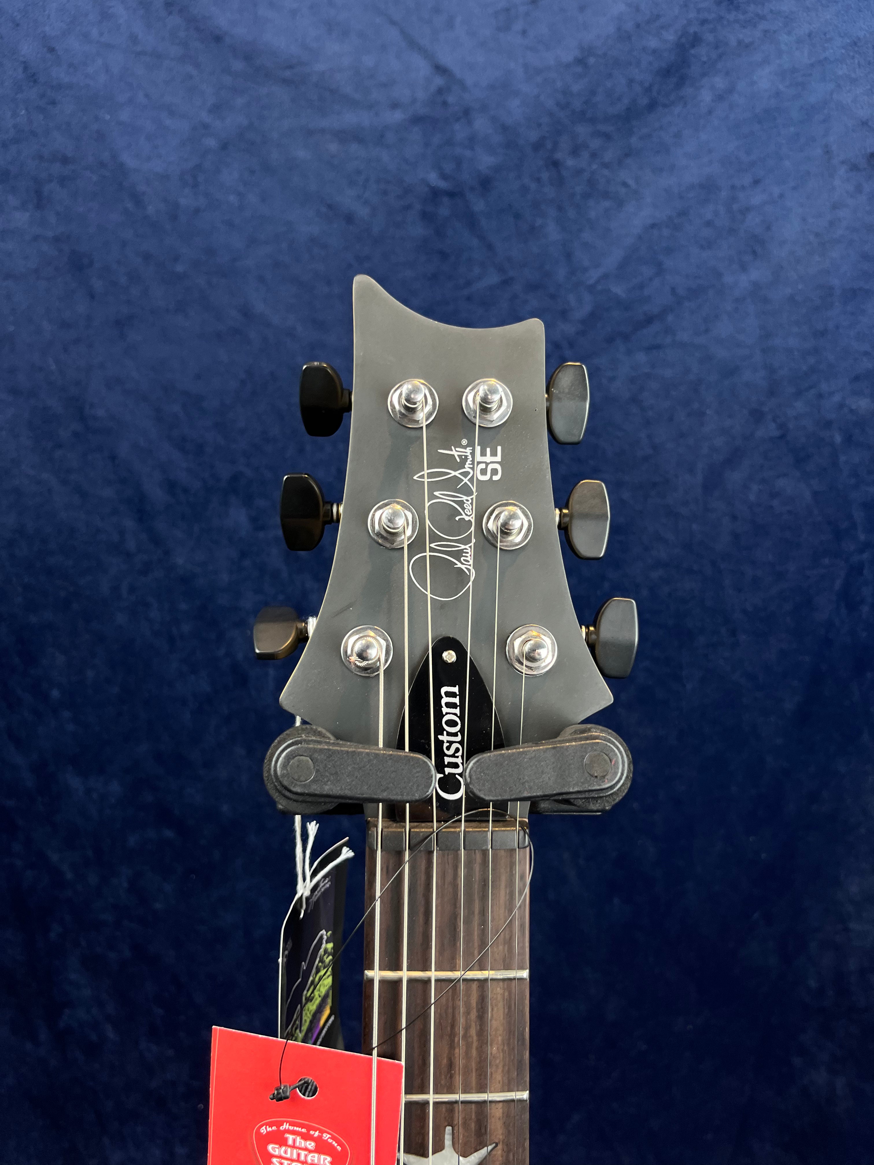 Six tuning pegs on a PRS SE electric guitar headstock, black finish, custom truss rod cover, with tags hanging from the neck against a blue background.