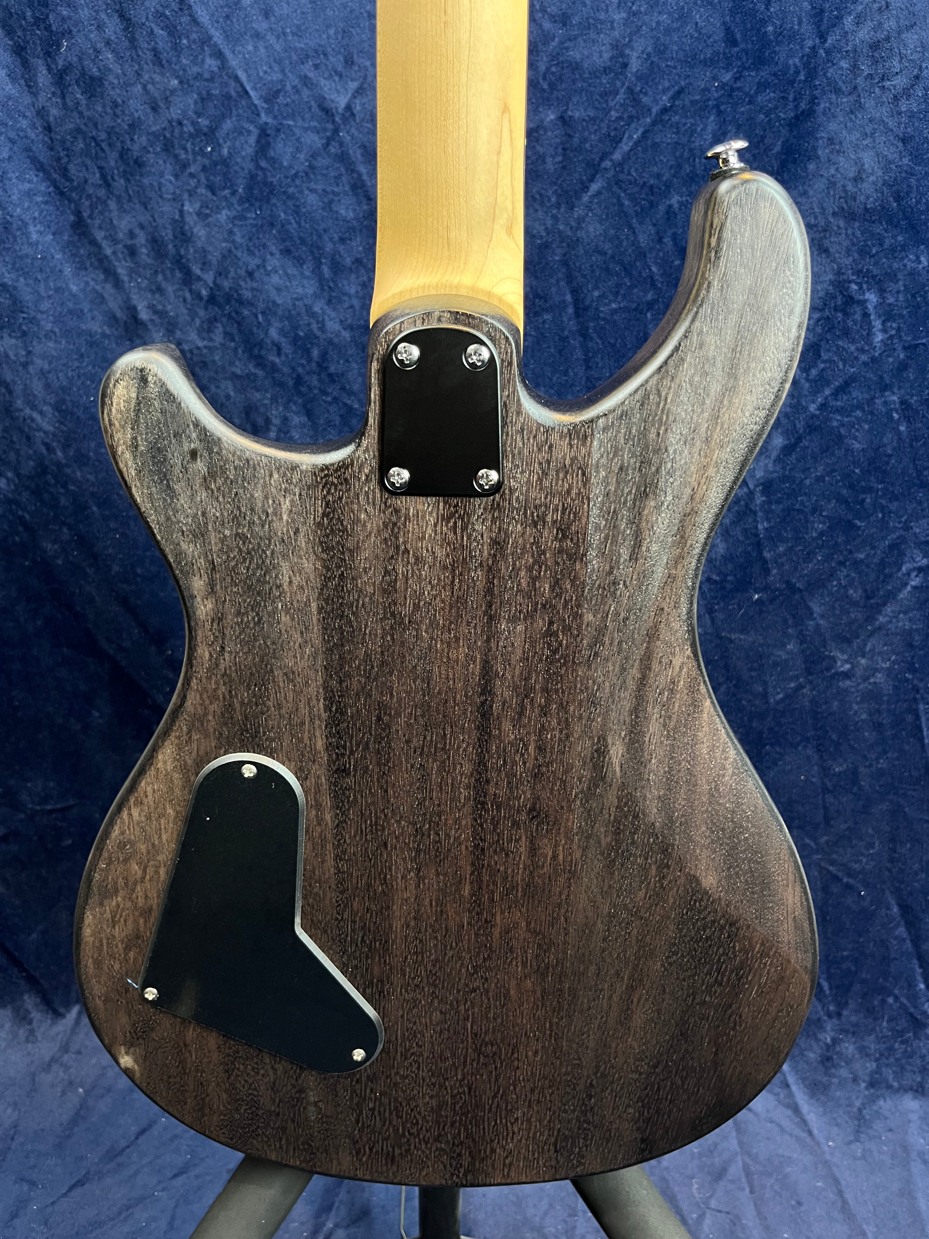 Back of an electric guitar with a dark wood grain finish, black control cavity covers, and a light-colored neck against a blue background.