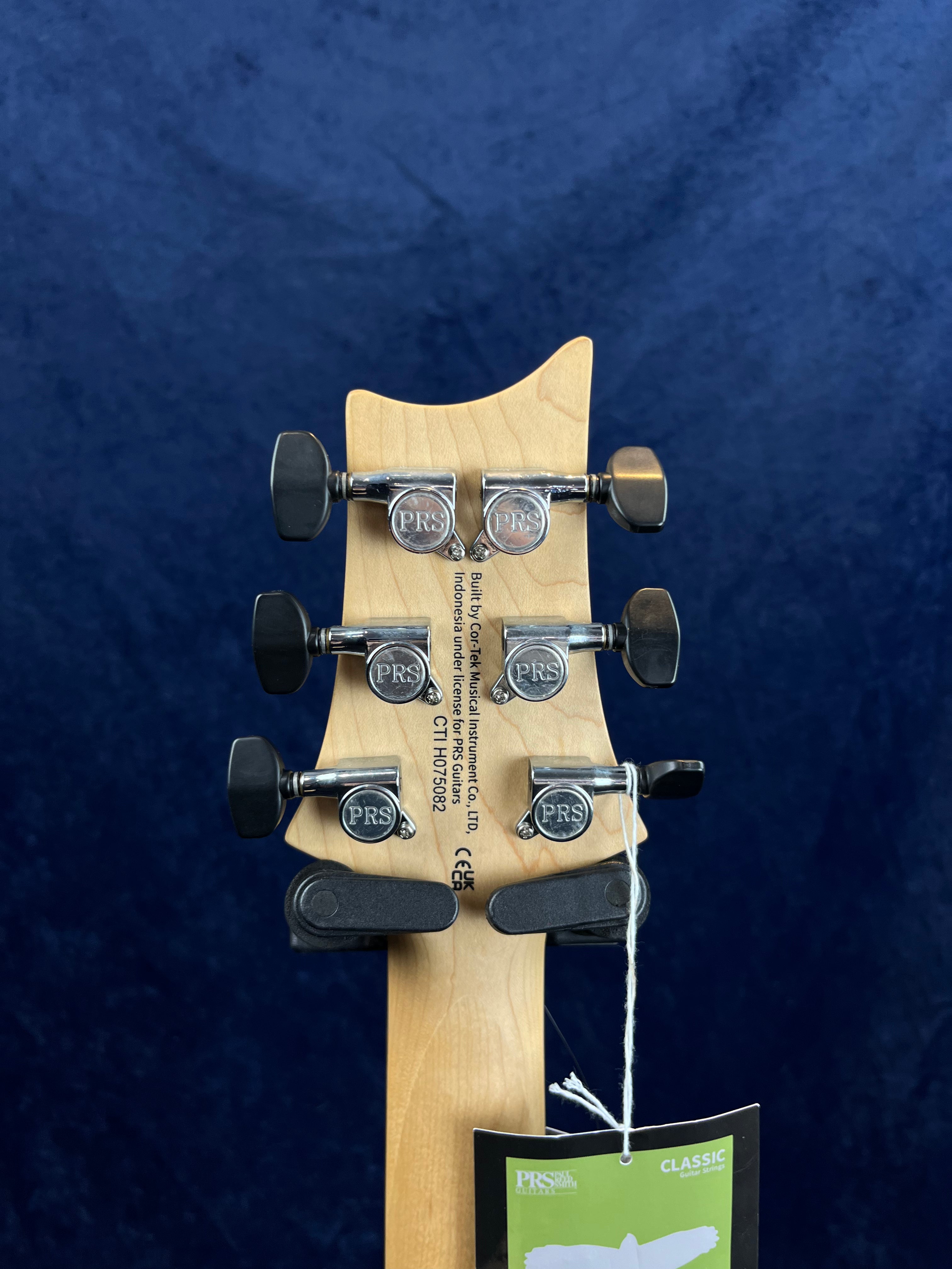 Back of a PRS electric guitar headstock with six chrome tuning pegs, black tuner buttons, serial number, and a green PRS Classic tag hanging.