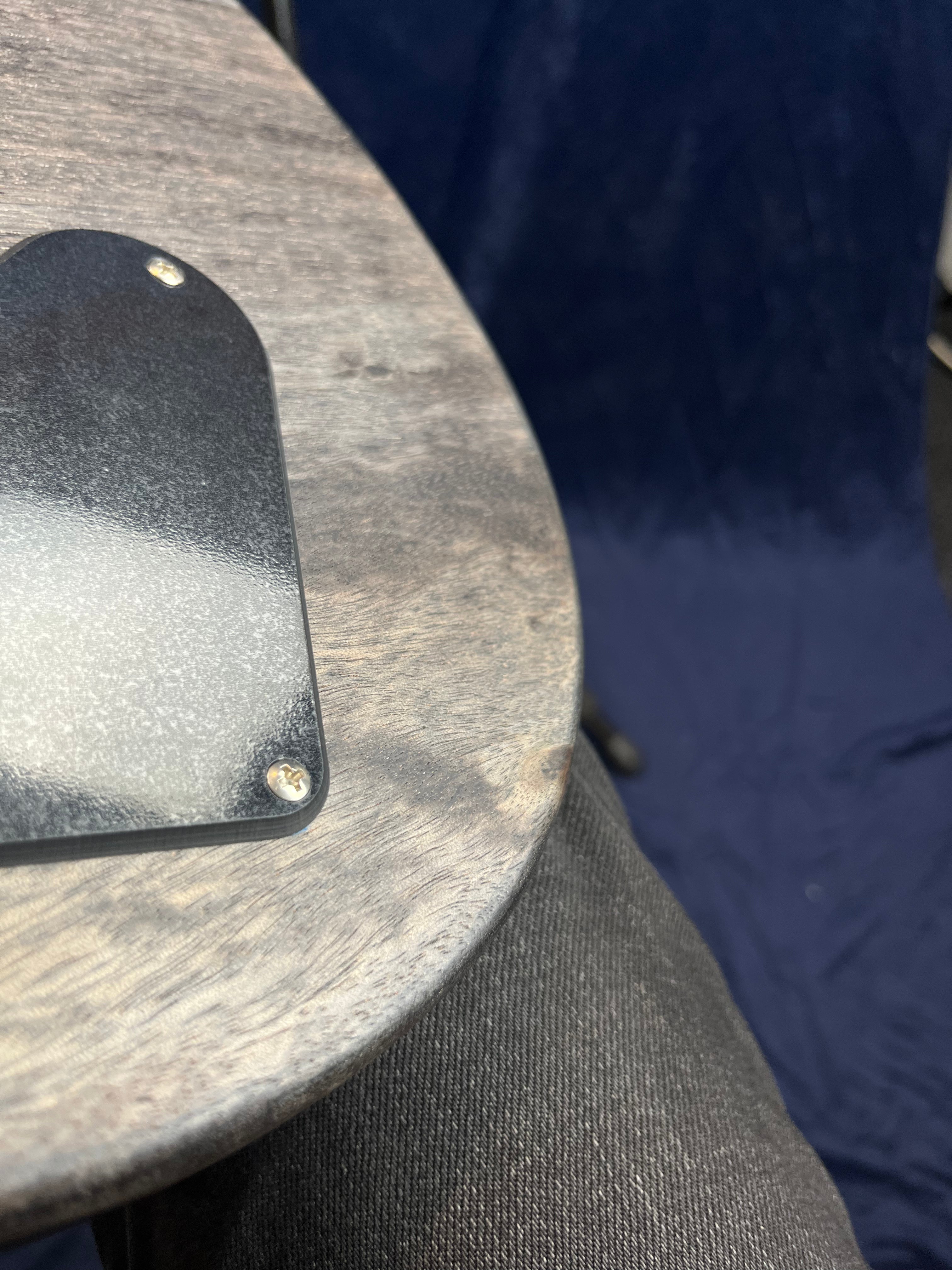 Close-up of the back of an electric guitar in natural wood finish, showing a black plastic cover plate with two screws. Denim-clad leg is visible below.