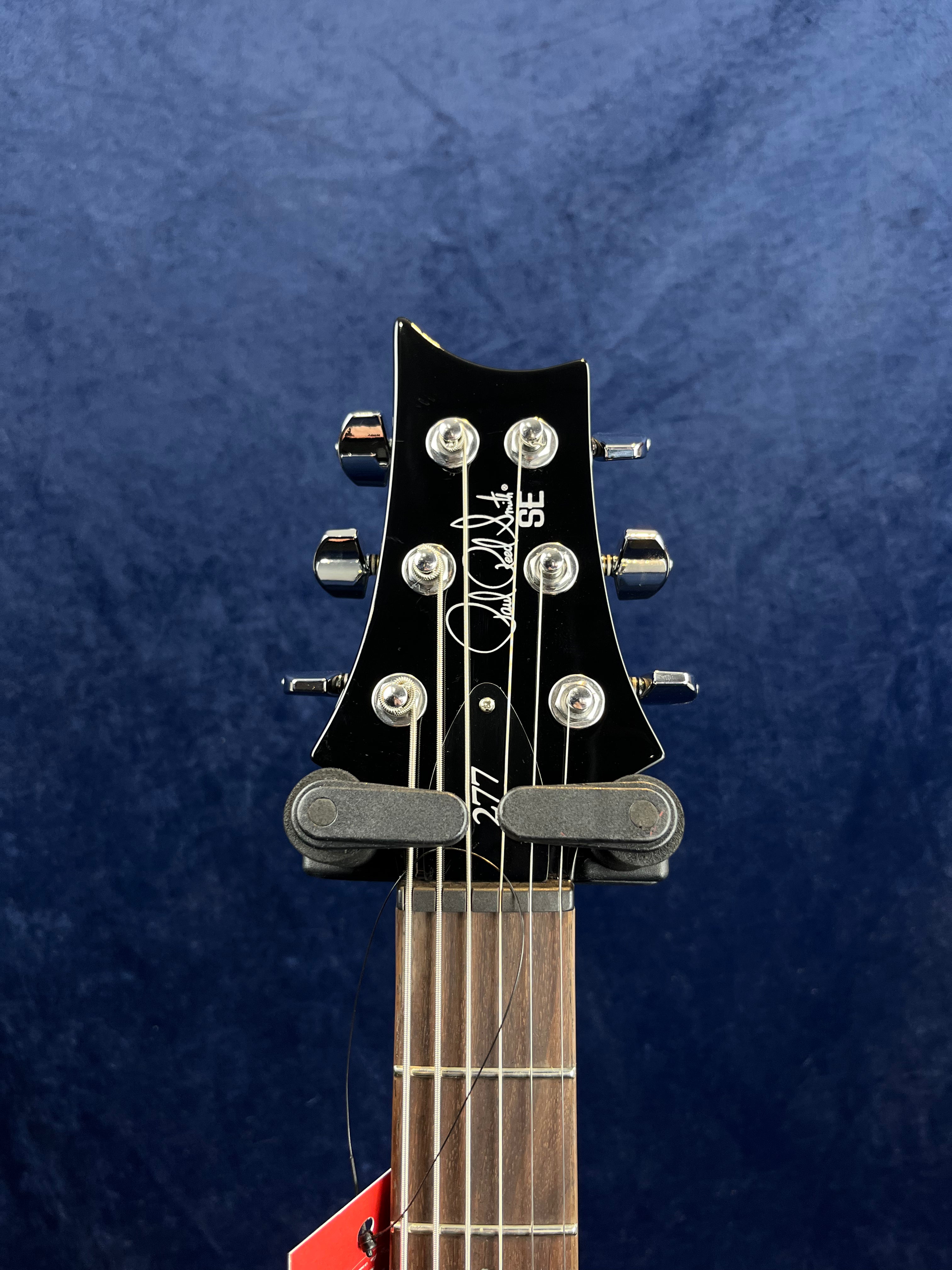 Electric guitar headstock with six tuning pegs, black finish, signature and "SE" logo on the front, part of a red tag visible near the fretboard.