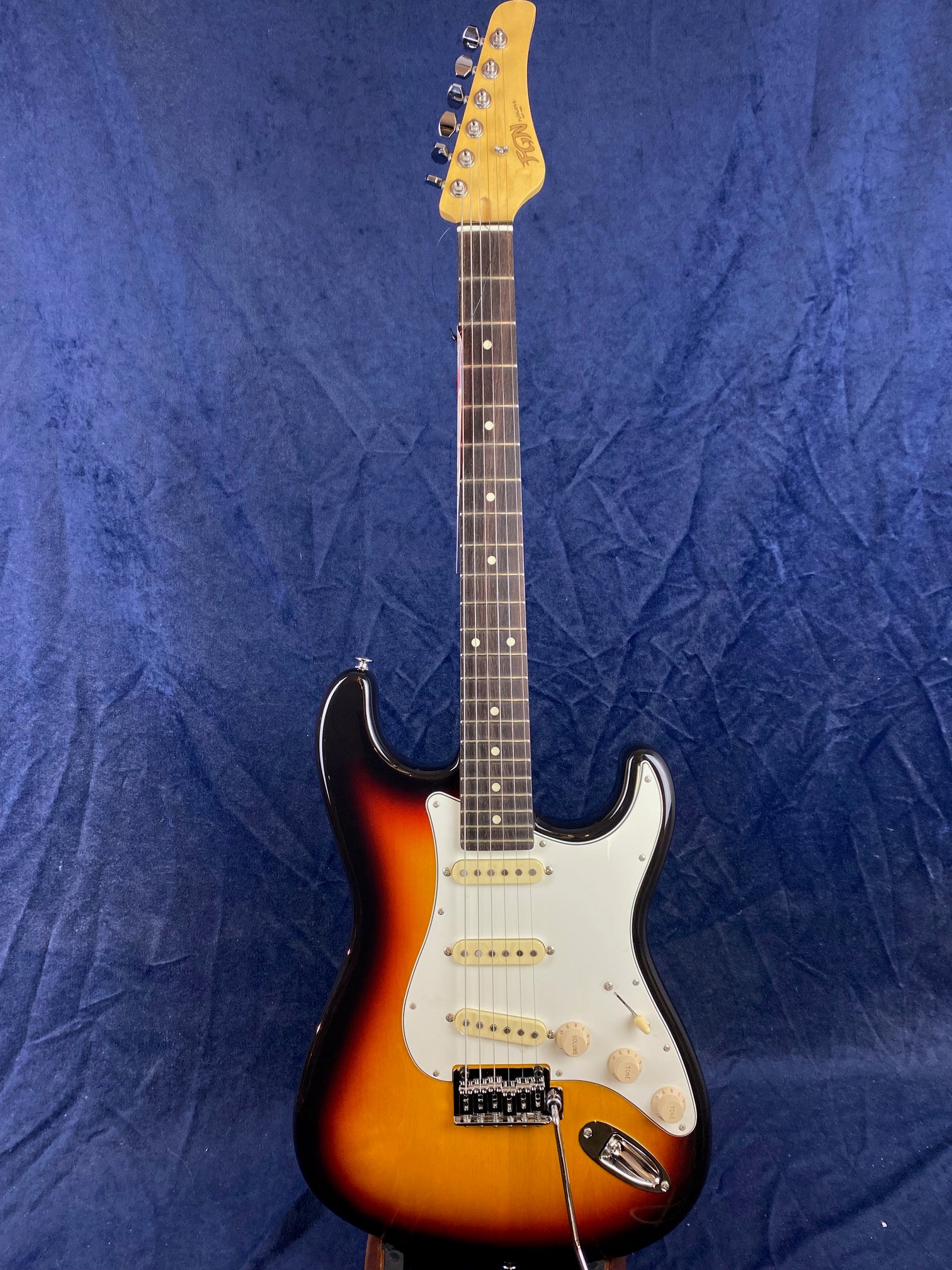 Sunburst electric guitar with a white pickguard, three single-coil pickups, and six tuning pegs against a dark blue background.