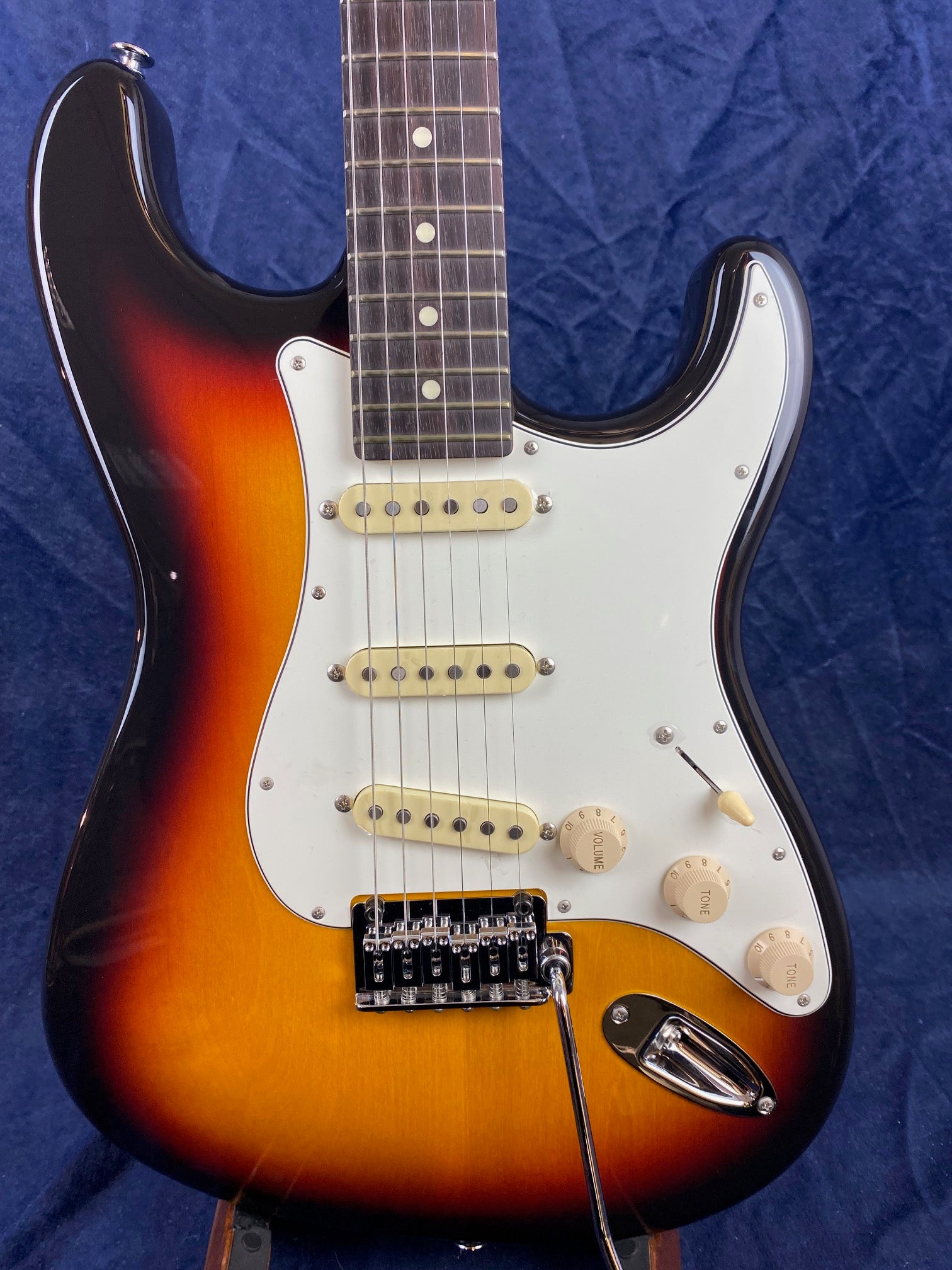 Sunburst electric guitar with a white pickguard, three single-coil pickups, three control knobs, and a whammy bar against a dark blue background.
