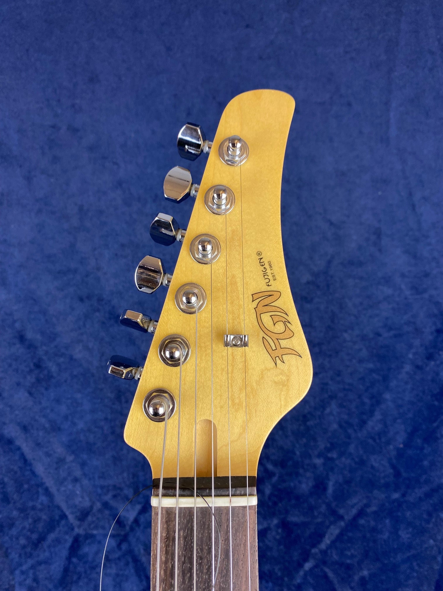 Electric guitar headstock with six tuning pegs, light wood finish, and the brand name "Fender" in stylized script on a dark blue background.