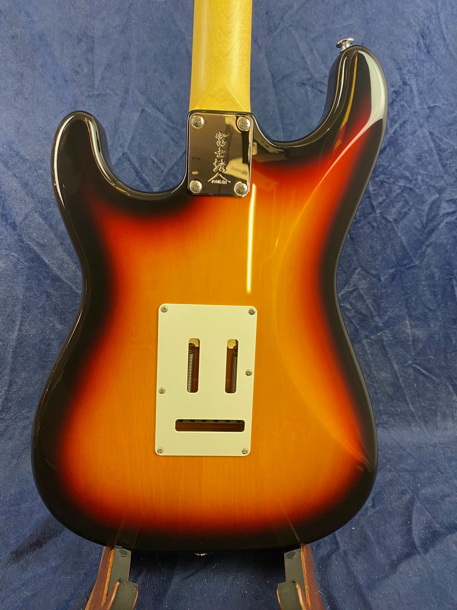 Sunburst electric guitar back view, with a rectangular white plate and visible tremolo spring slots, set against a dark blue textured background.