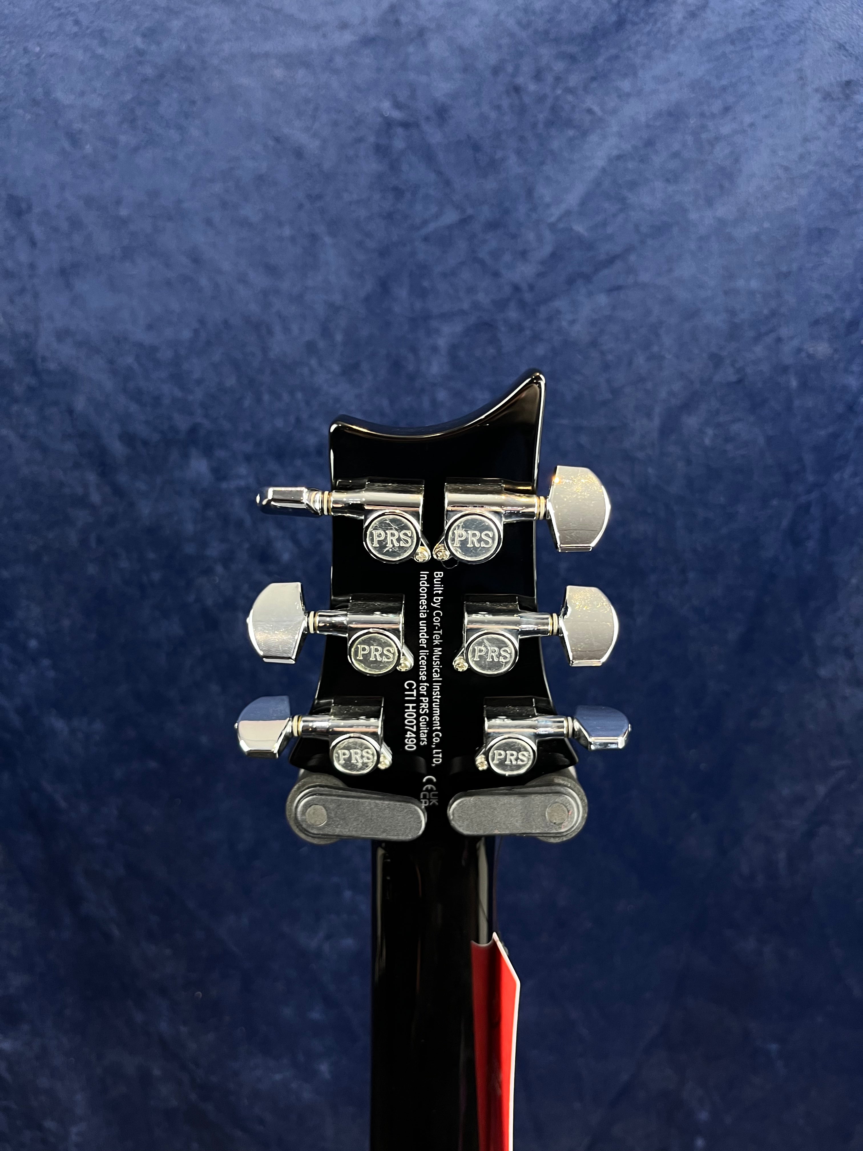 Back of a PRS electric guitar headstock with six chrome tuning pegs, PRS logo on each peg, against a dark blue background.