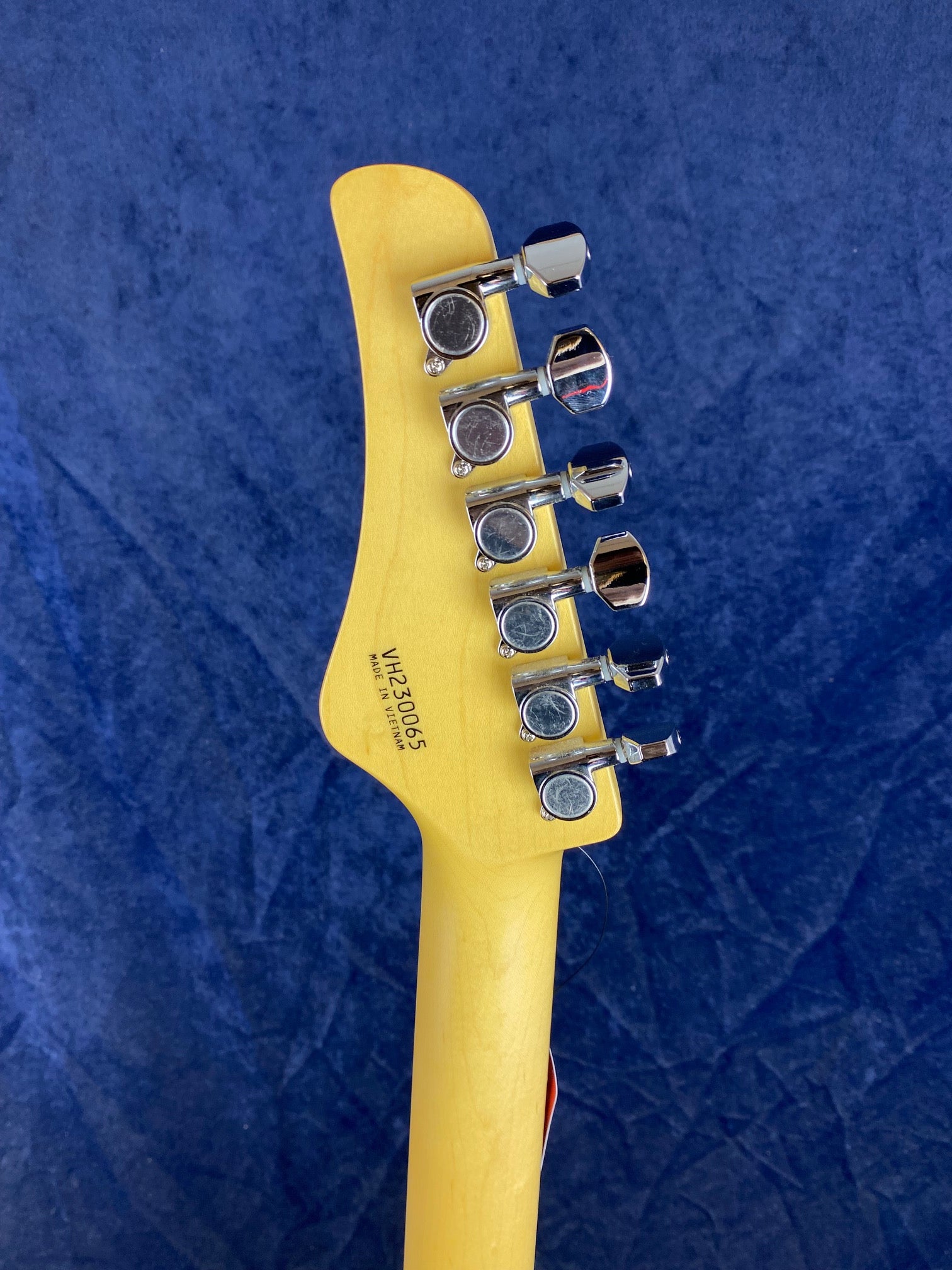 Six chrome tuning pegs line the back of a light wood electric guitar headstock against a blue background, with a serial number and "Made in Vietnam" visible.
