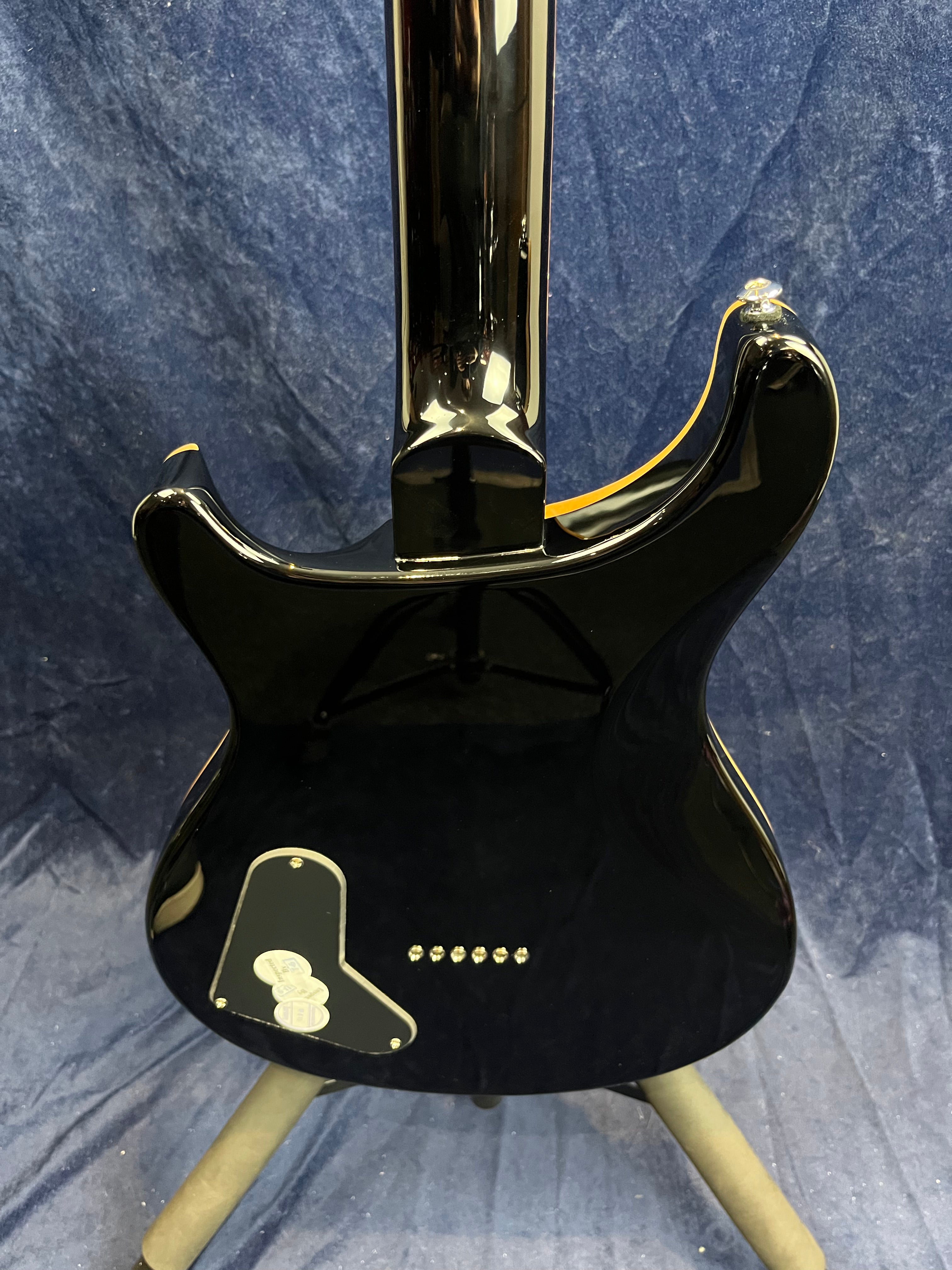 Glossy black electric guitar seen from the back, with a rectangular electronics cover and six string ferrules in a row on the body.