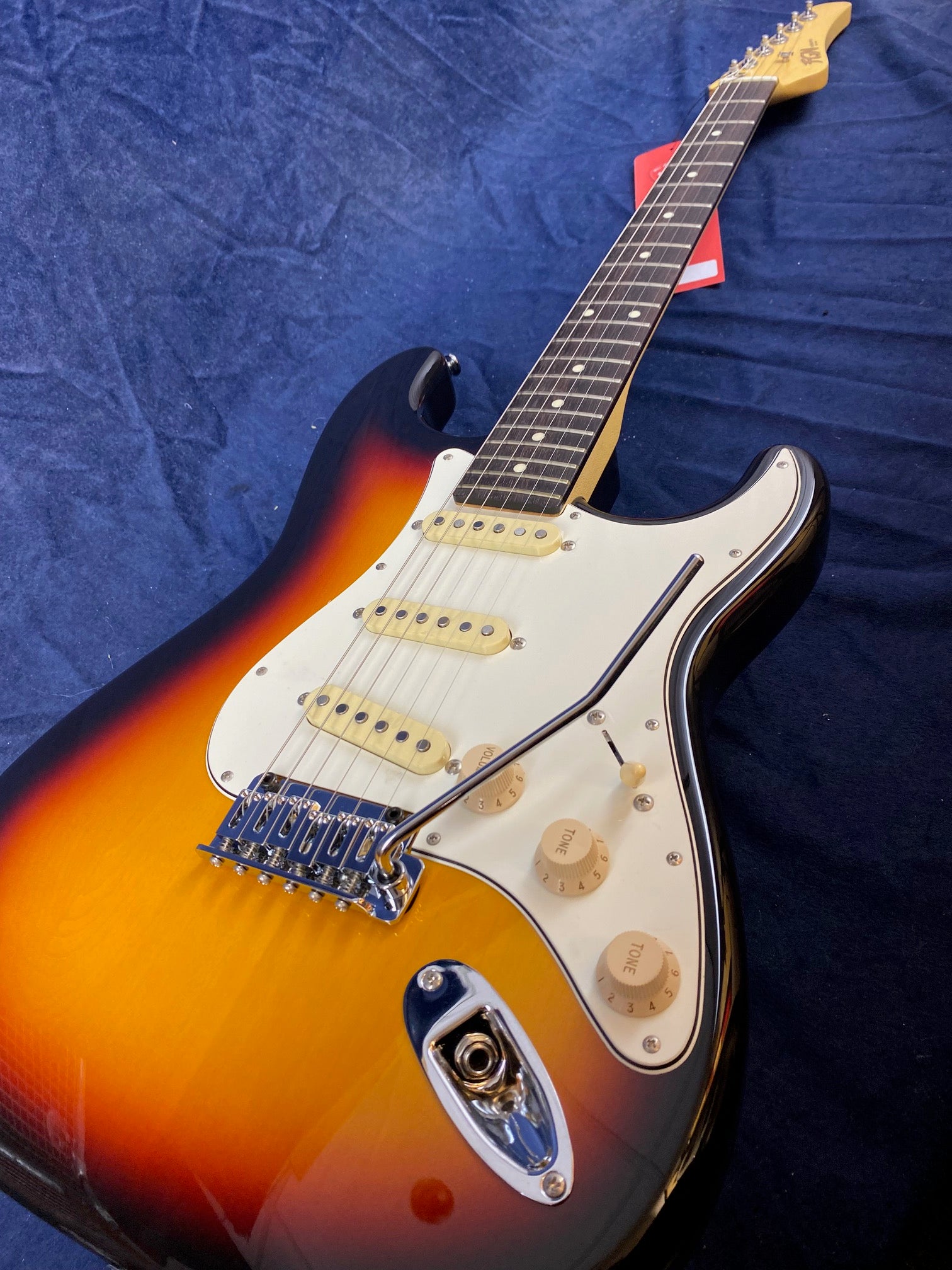Electric guitar with a sunburst finish, white pickguard, three single-coil pickups, three control knobs, and a whammy bar lying on a dark cloth.