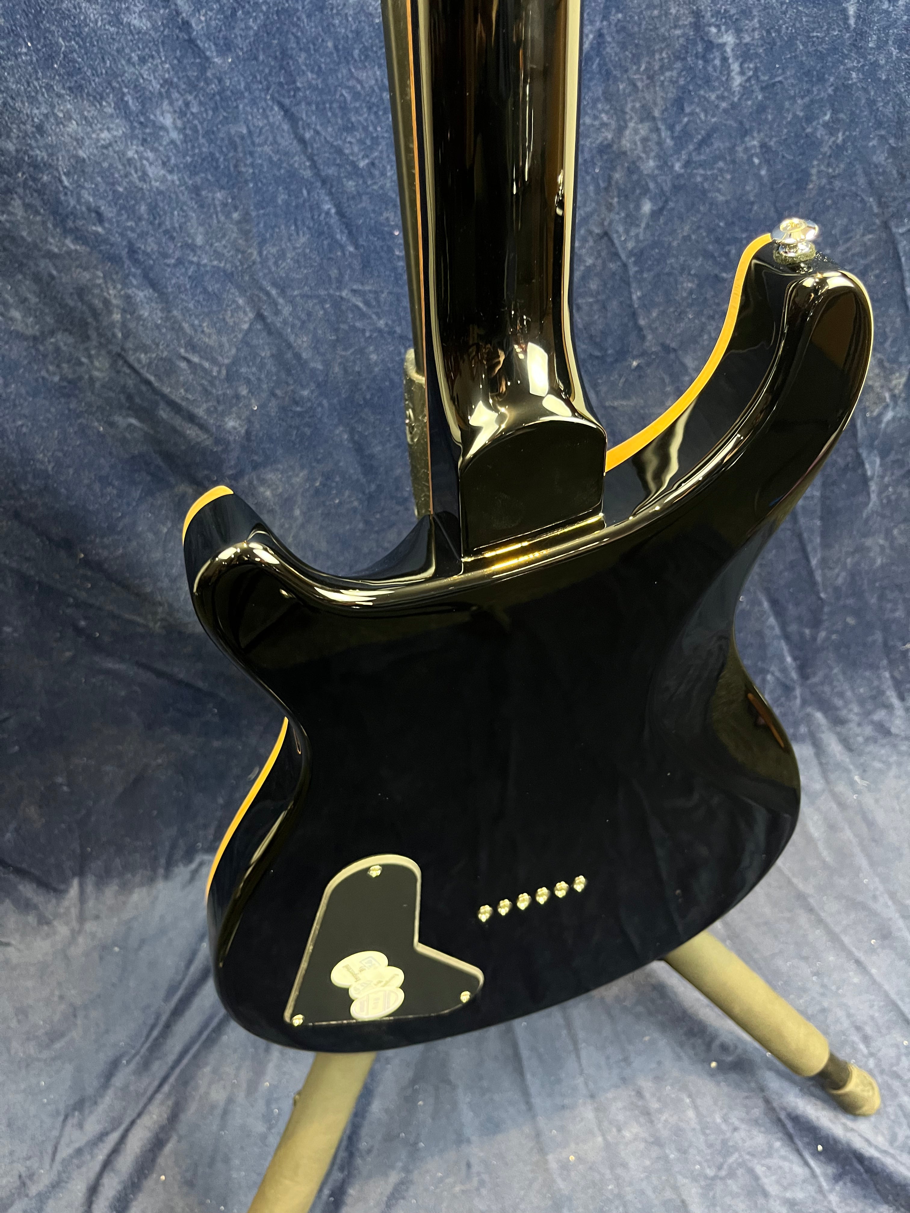 Glossy black electric guitar viewed from the back, yellow edge trim, six rear string ferrules, and a black plastic cover on the lower left side.