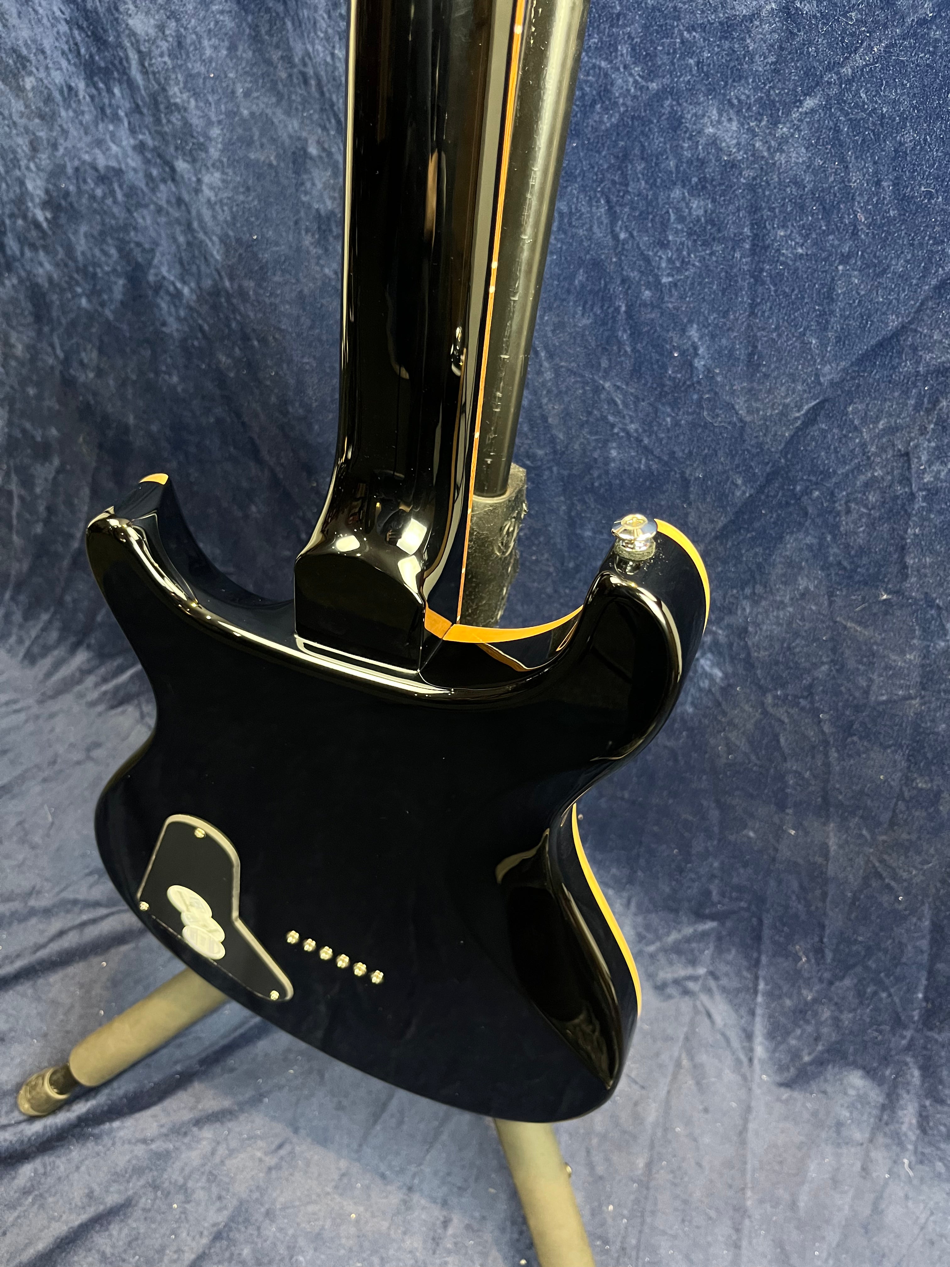 Glossy black electric guitar showing the back of the body and neck, with cream binding and a control cavity cover, resting on a stand against a blue background.