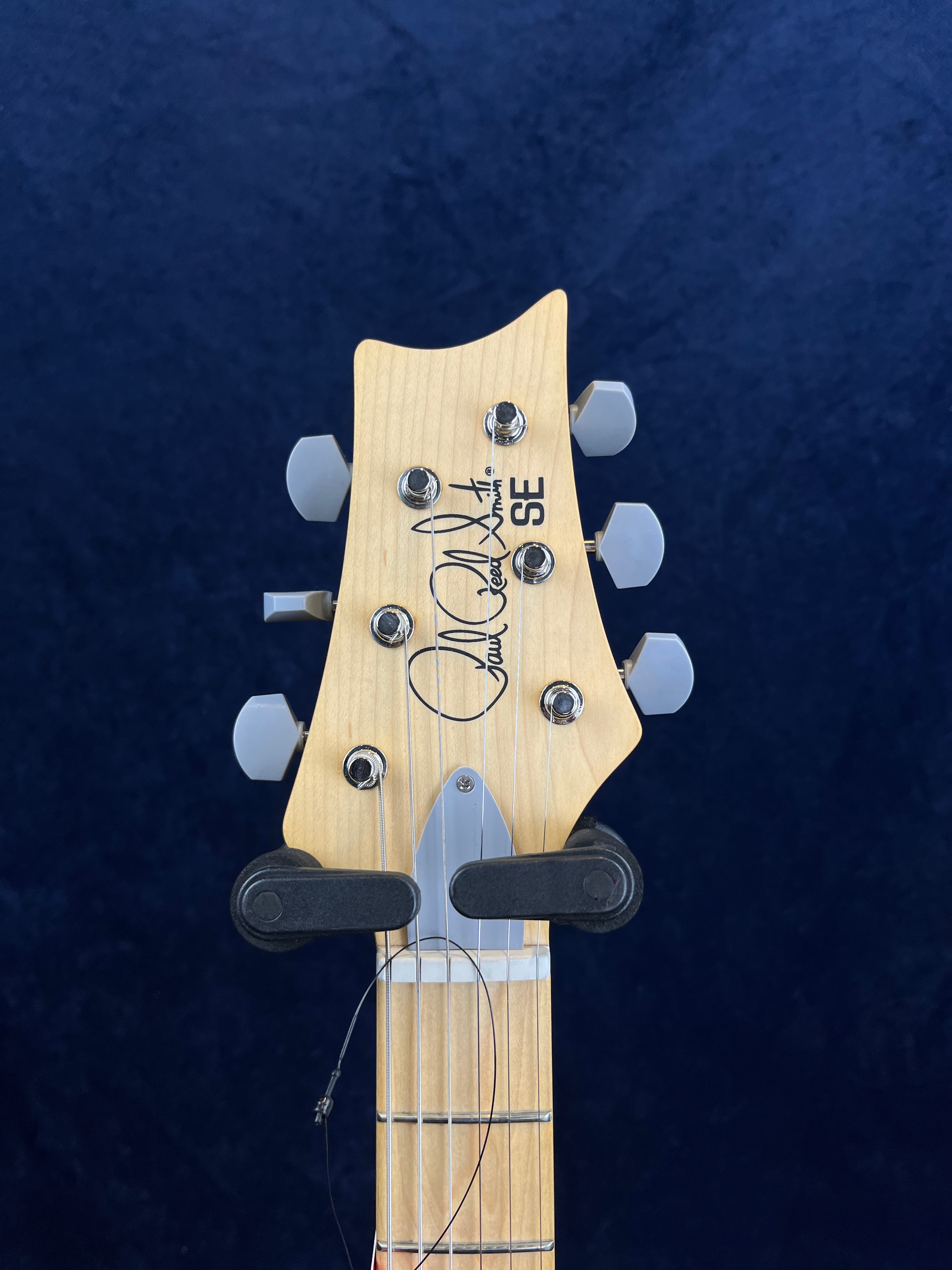 Light wood electric guitar headstock with six silver tuning pegs, signature, “SE,” and part of a hanging bracket visible.