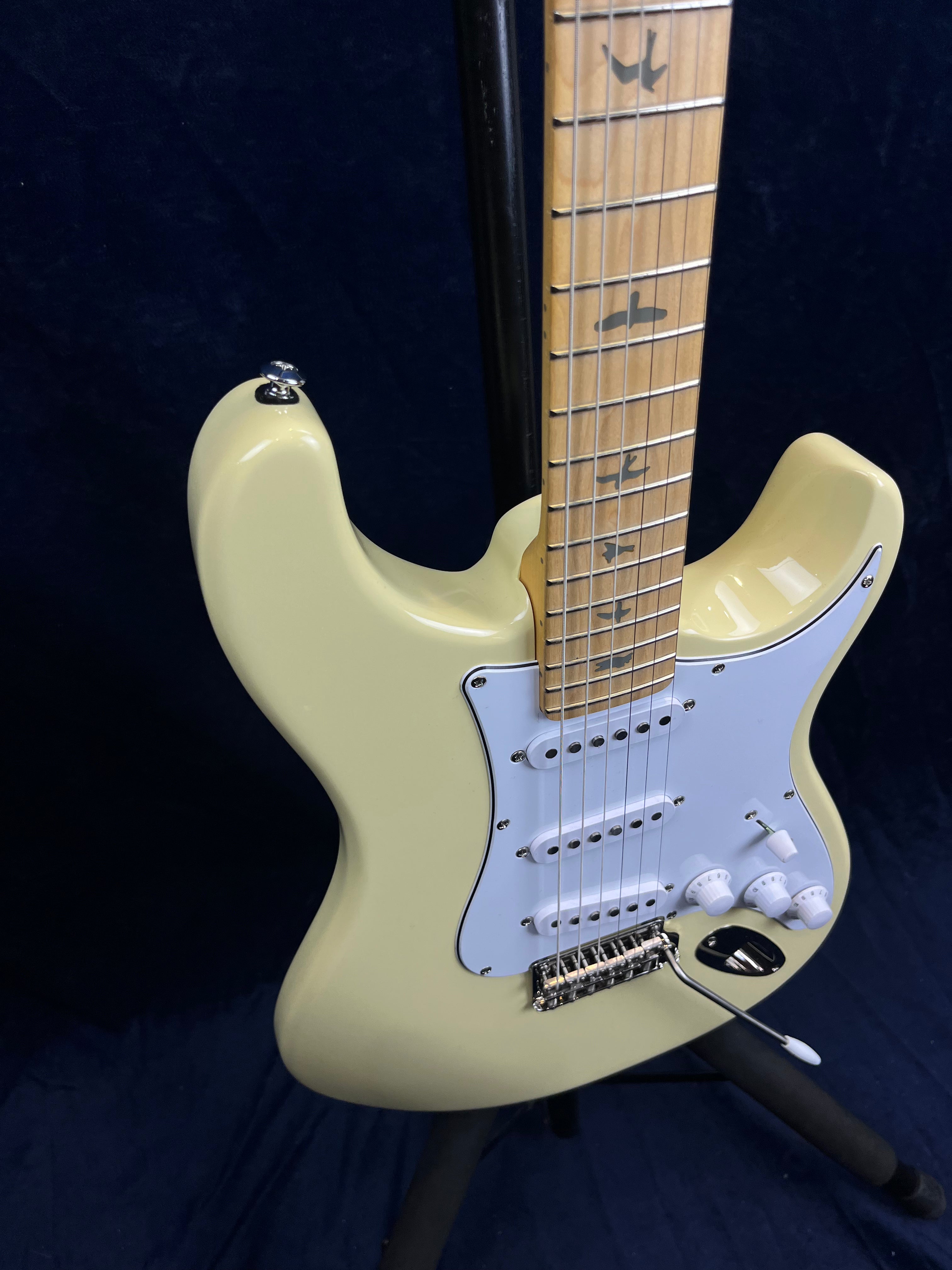 Cream-colored electric guitar with a white pickguard, three single-coil pickups, tremolo arm, and bird-shaped inlays on a light maple neck.
