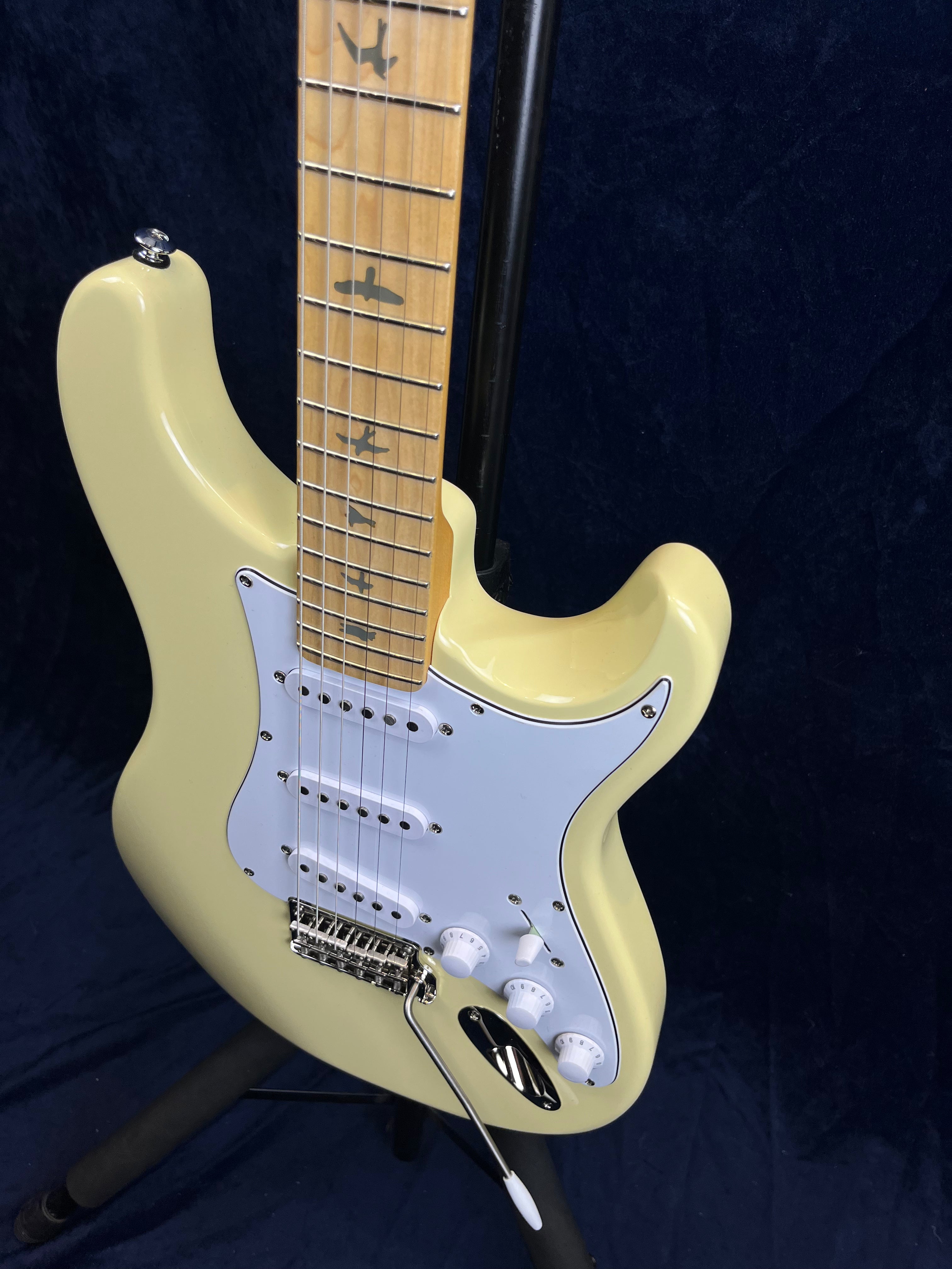 Cream-colored electric guitar with a light pickguard, three single-coil pickups, bird inlays on maple fretboard, and a whammy bar attached.