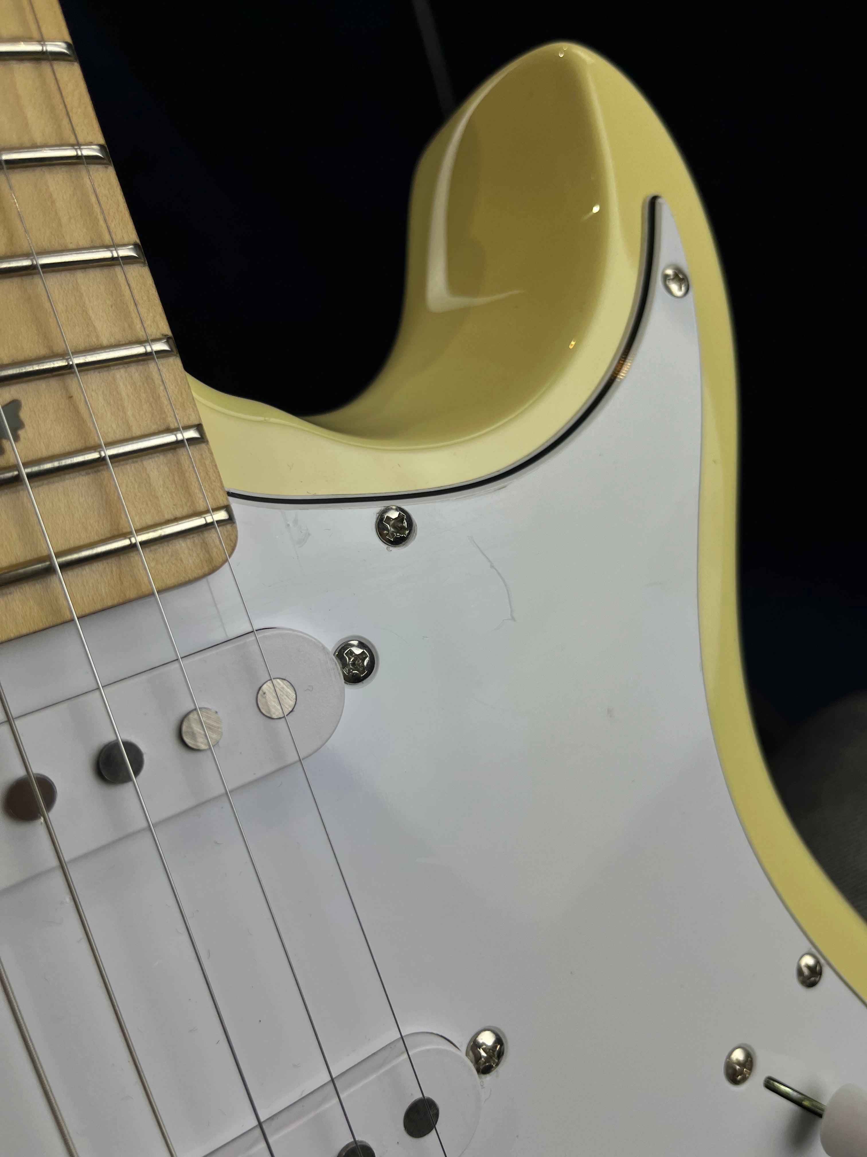 Close-up of a cream-colored electric guitar body showing strings, pickups, pickguard screws, and part of the maple neck with metal frets.