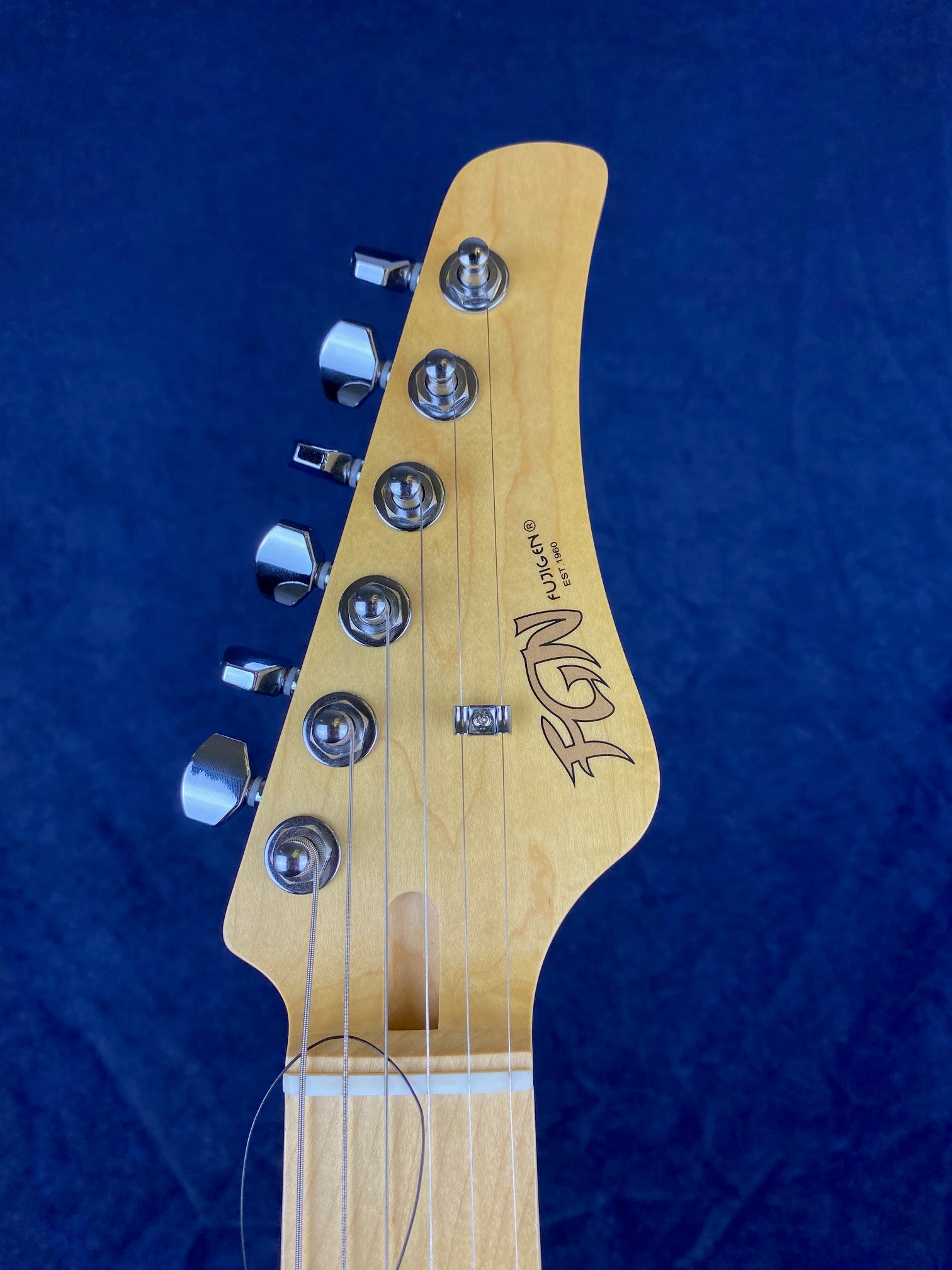 Light wood electric guitar headstock with six tuning pegs, metal strings, and the "FüLGEN" brand logo on a dark blue background.