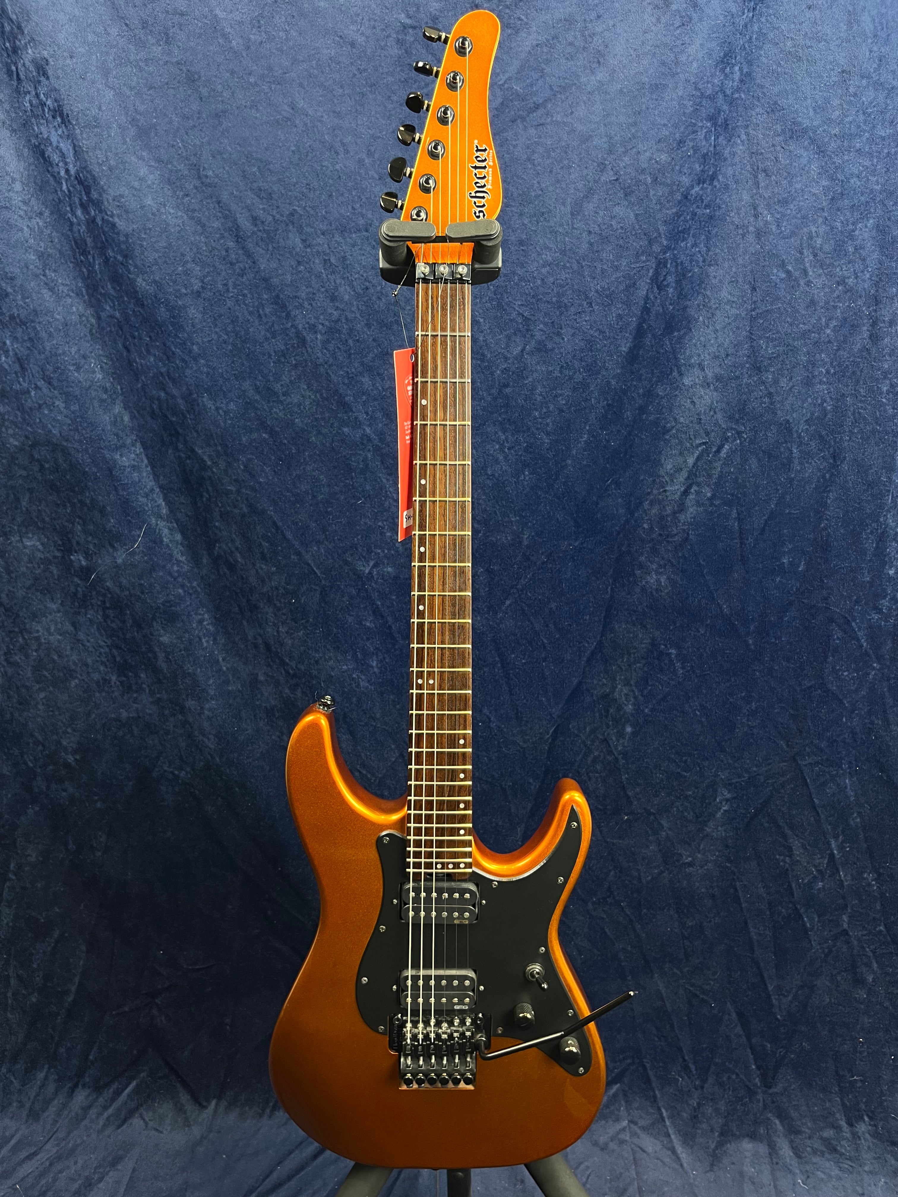 Metallic orange Schecter electric guitar with double cutaway body, black pickguard, two humbucker pickups, and a whammy bar against a dark backdrop.