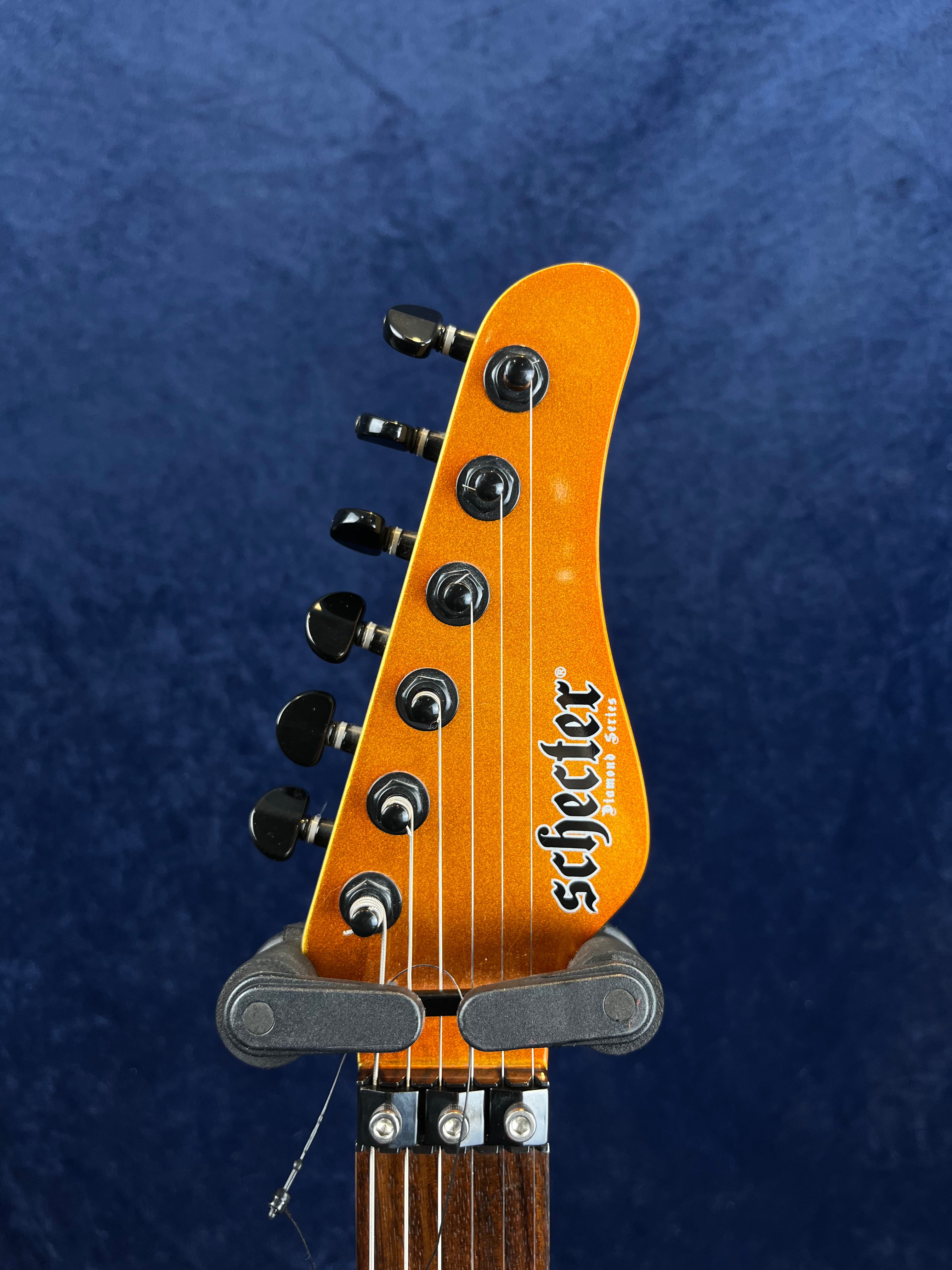 Orange Schecter electric guitar headstock with six black tuning pegs, "Diamond Series" label, and locking nuts against a dark blue background.