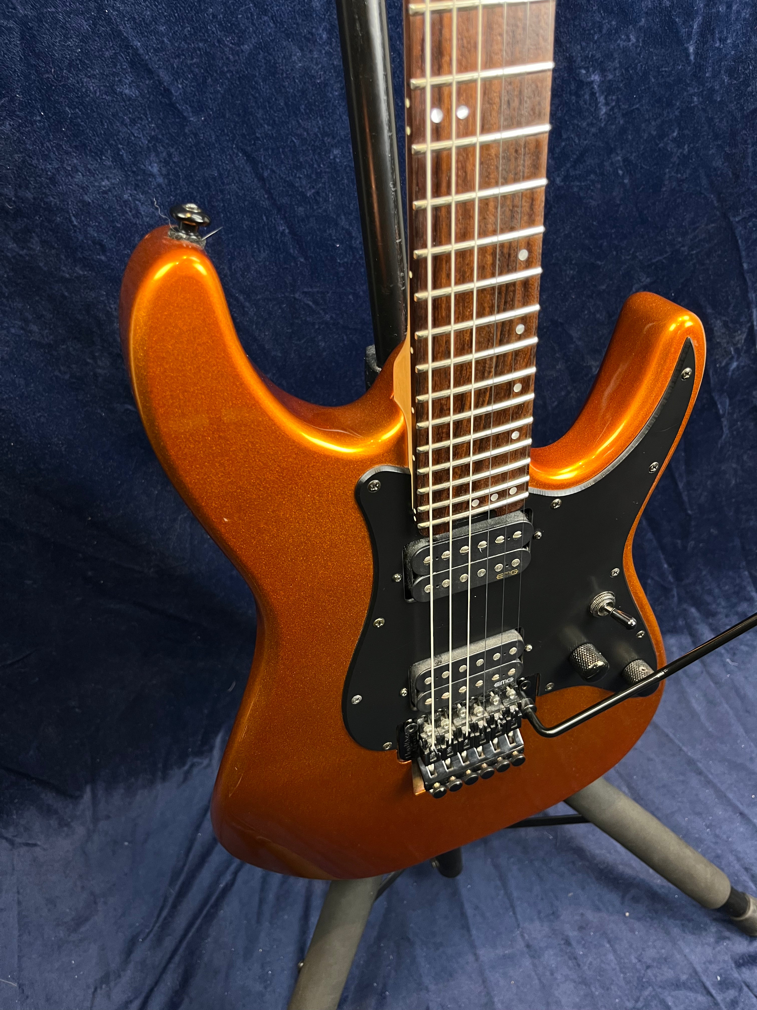 Metallic orange electric guitar with black pickguard, two humbucker pickups, a whammy bar, and a rosewood fretboard, resting on a stand.