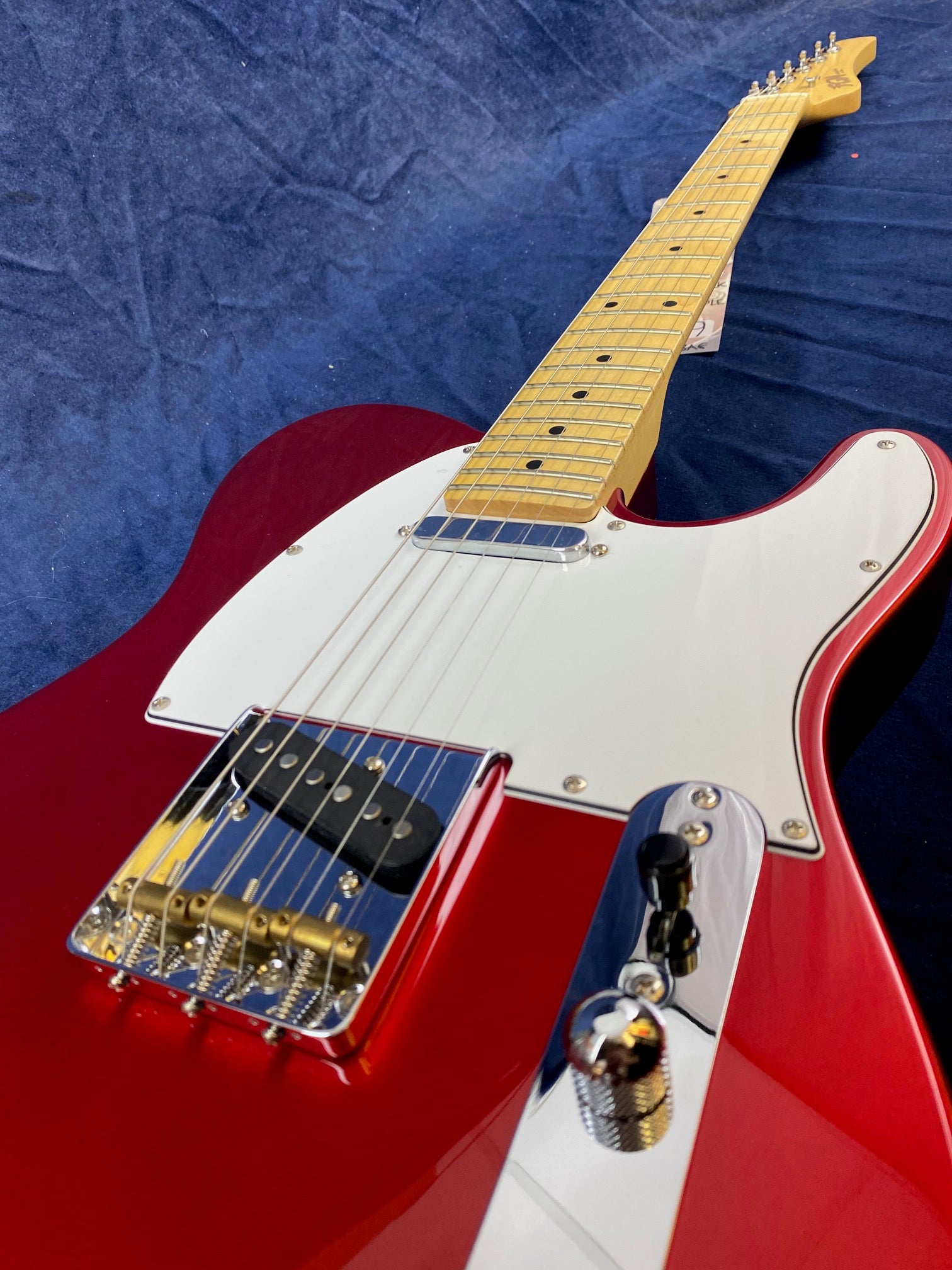 Red electric guitar with a white pickguard, chrome hardware, two pickups, control knobs, and a maple neck lying on a dark blue surface.