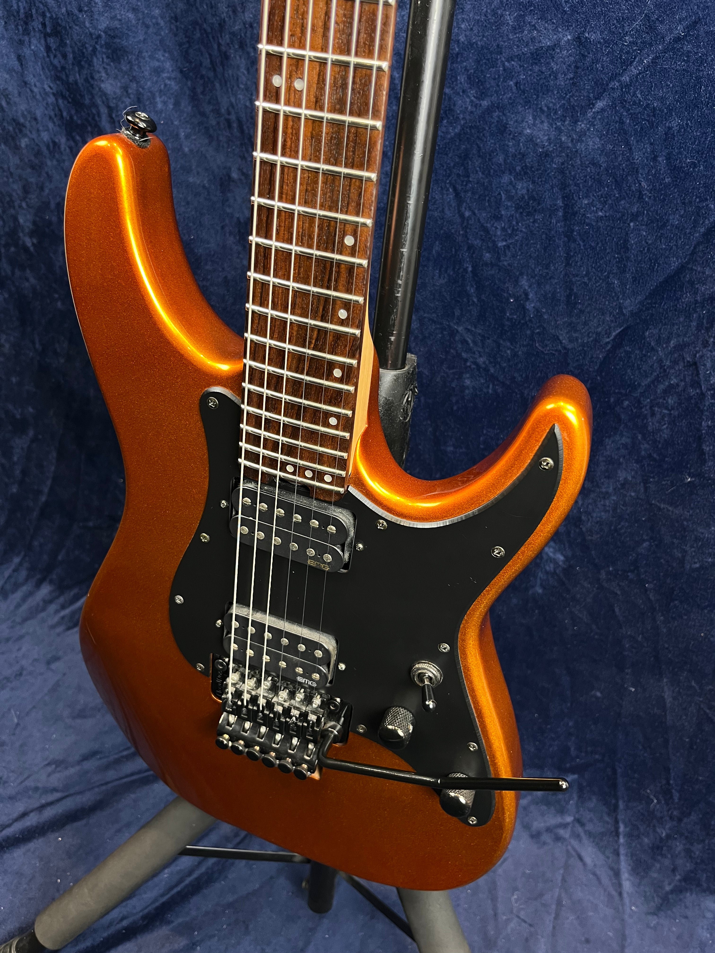 Metallic orange electric guitar with a black pickguard, dual humbucker pickups, a tremolo bridge, and a rosewood fretboard on a stand.