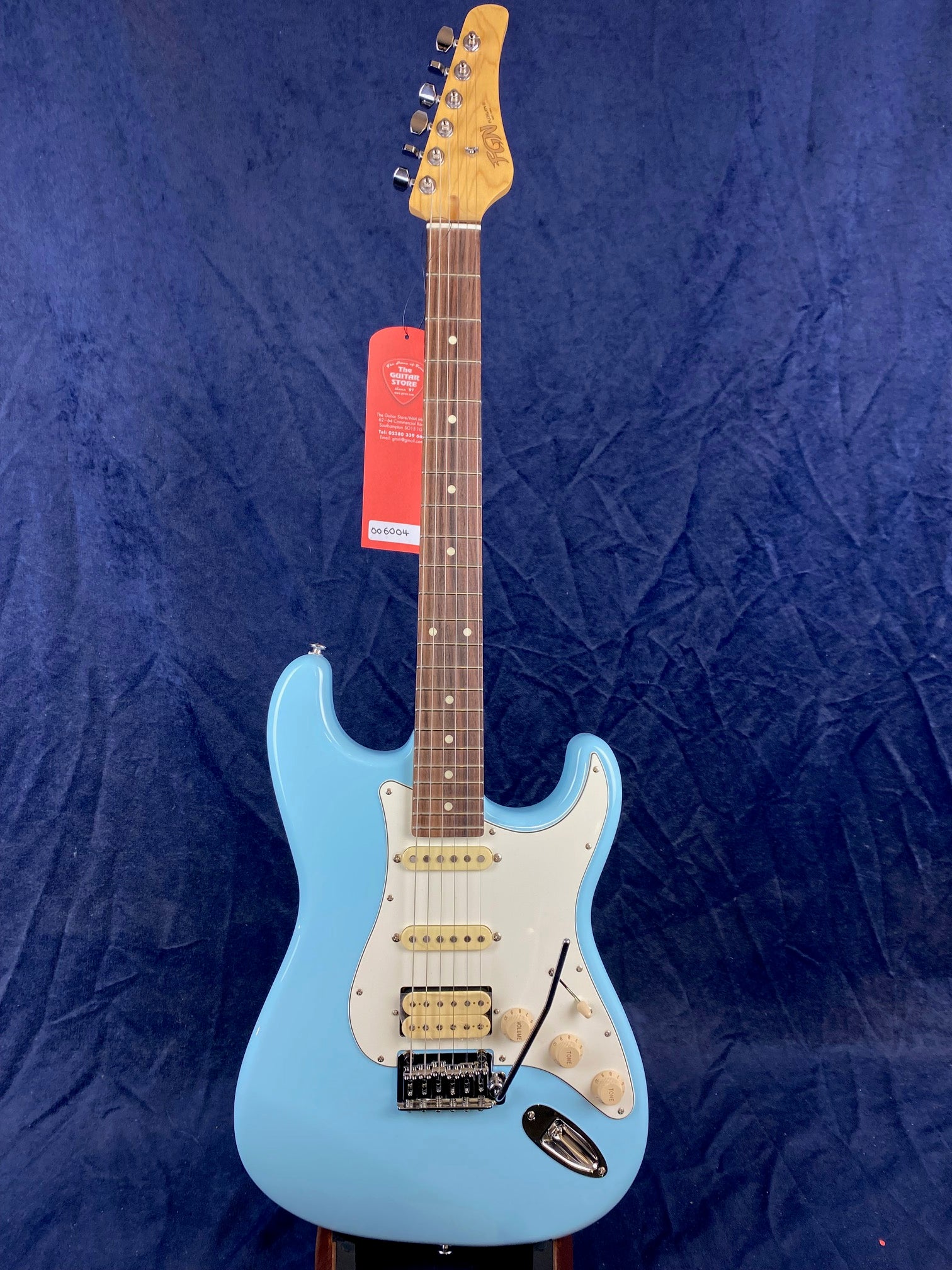 Light blue electric guitar with a white pickguard, three pickups, a tremolo bridge, and a maple neck, standing upright against a dark blue background.