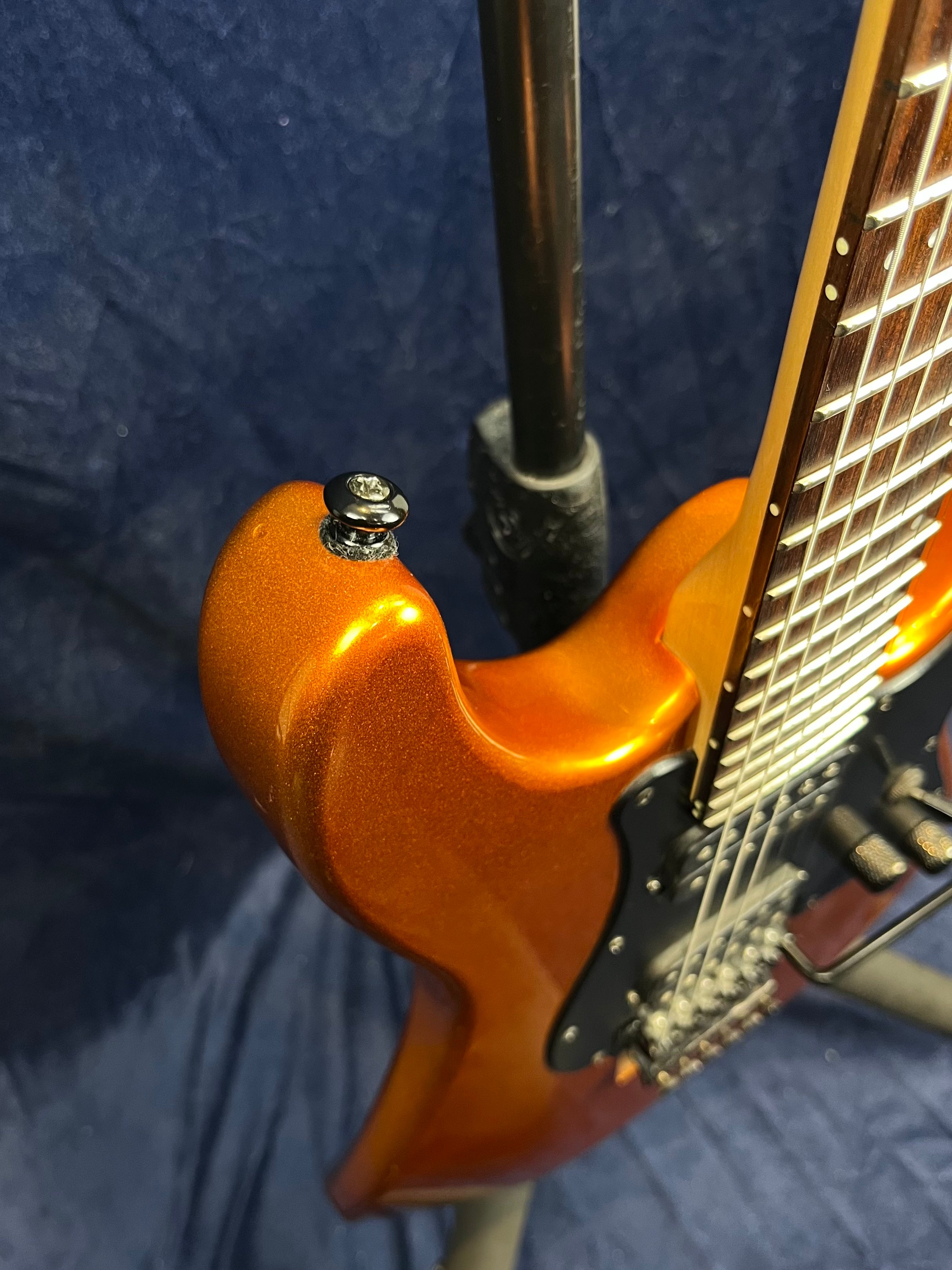 Metallic orange electric guitar showing upper body, strap button, black pickguard, part of fretboard with dot inlays, and neck on a stand.