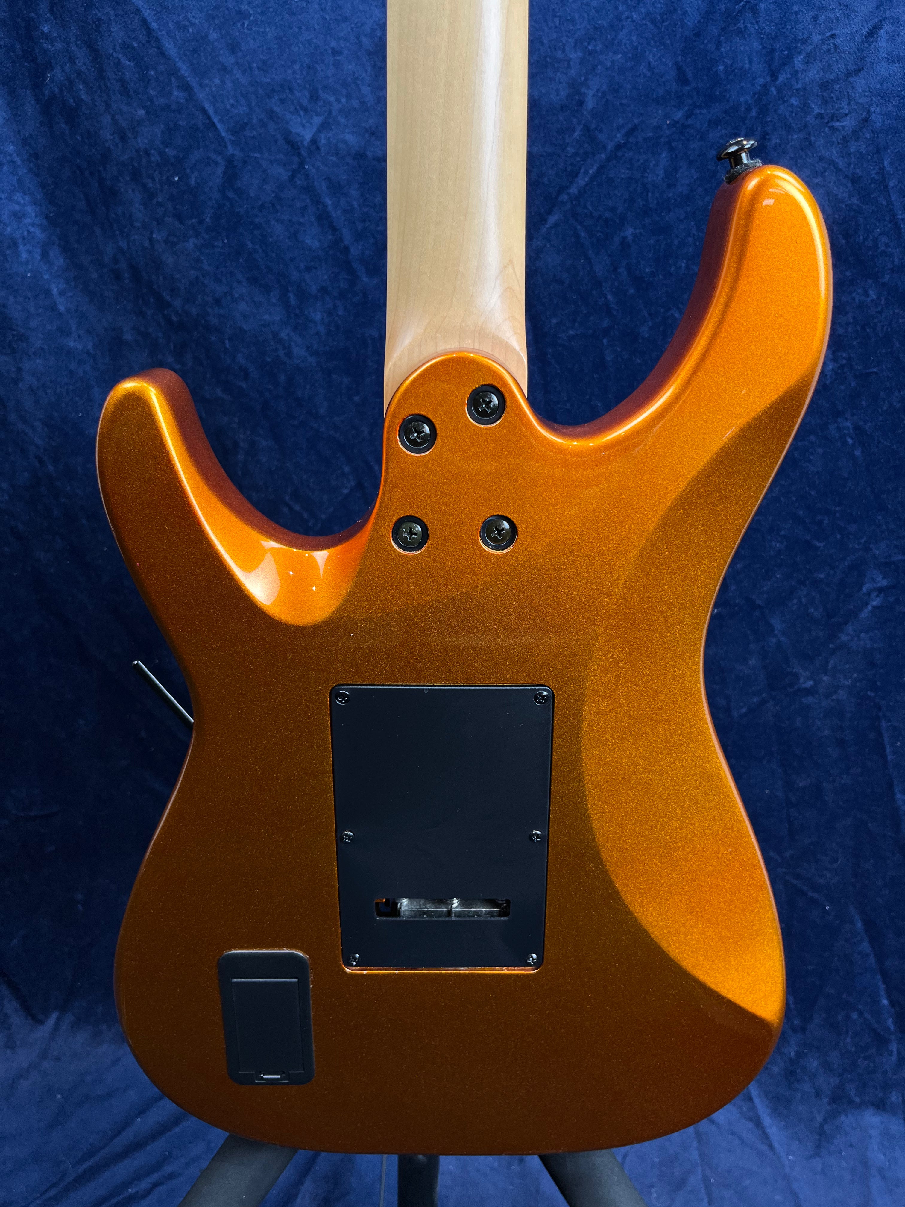 Electric guitar back view, metallic orange finish, four neck bolts, two black rear cavity covers, natural wood neck against a dark blue background.